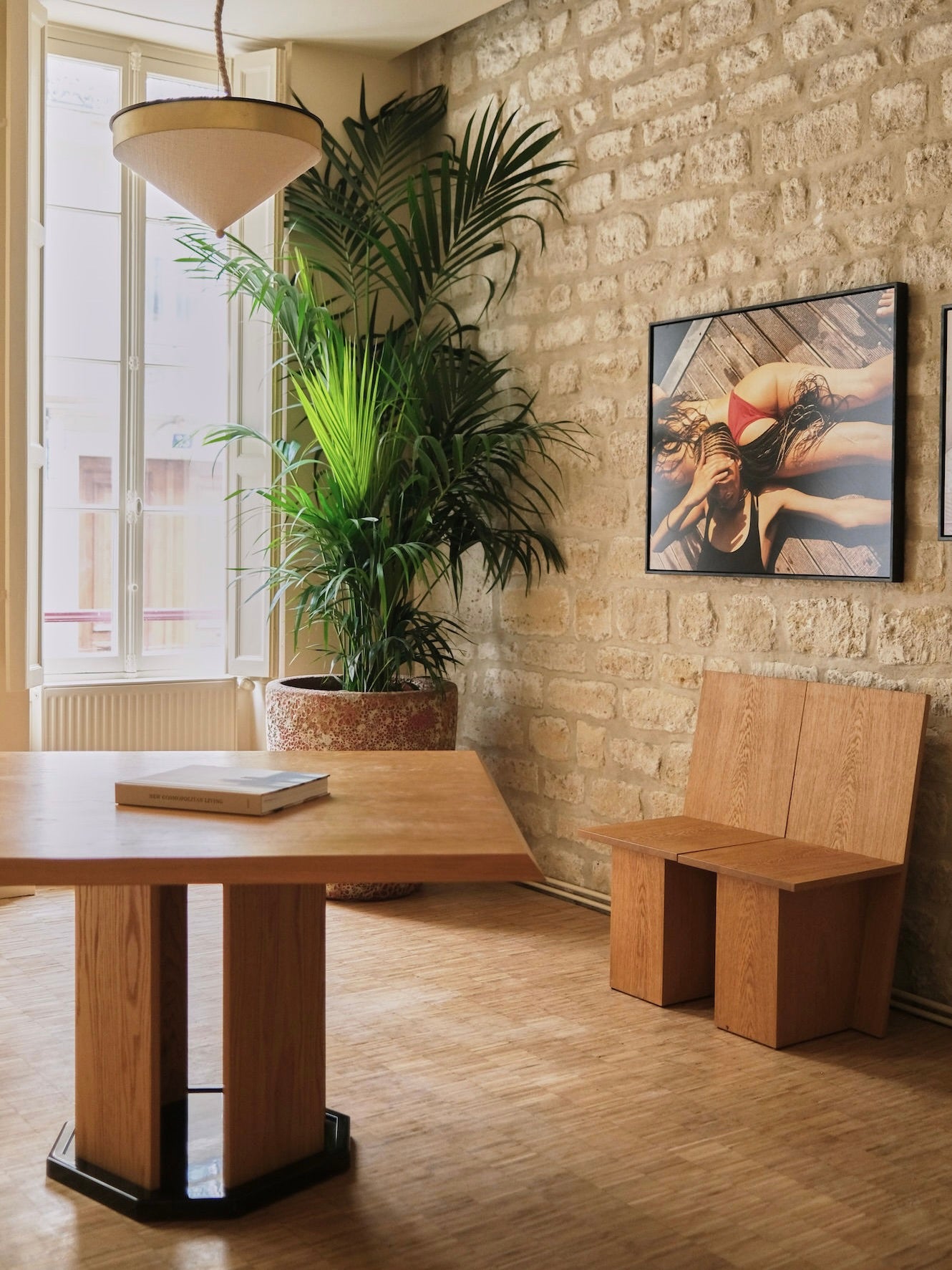 A stylish room showcases HAUVETTE & MADANI's "La Coupe" modular chair paired with a sleek wooden table, enhanced by the elegance of brushed oak. A large green plant flourishes near the window, while a wall is adorned with a framed photograph titled "La Coupe," capturing someone lounging in swimwear against stone and parquet flooring.