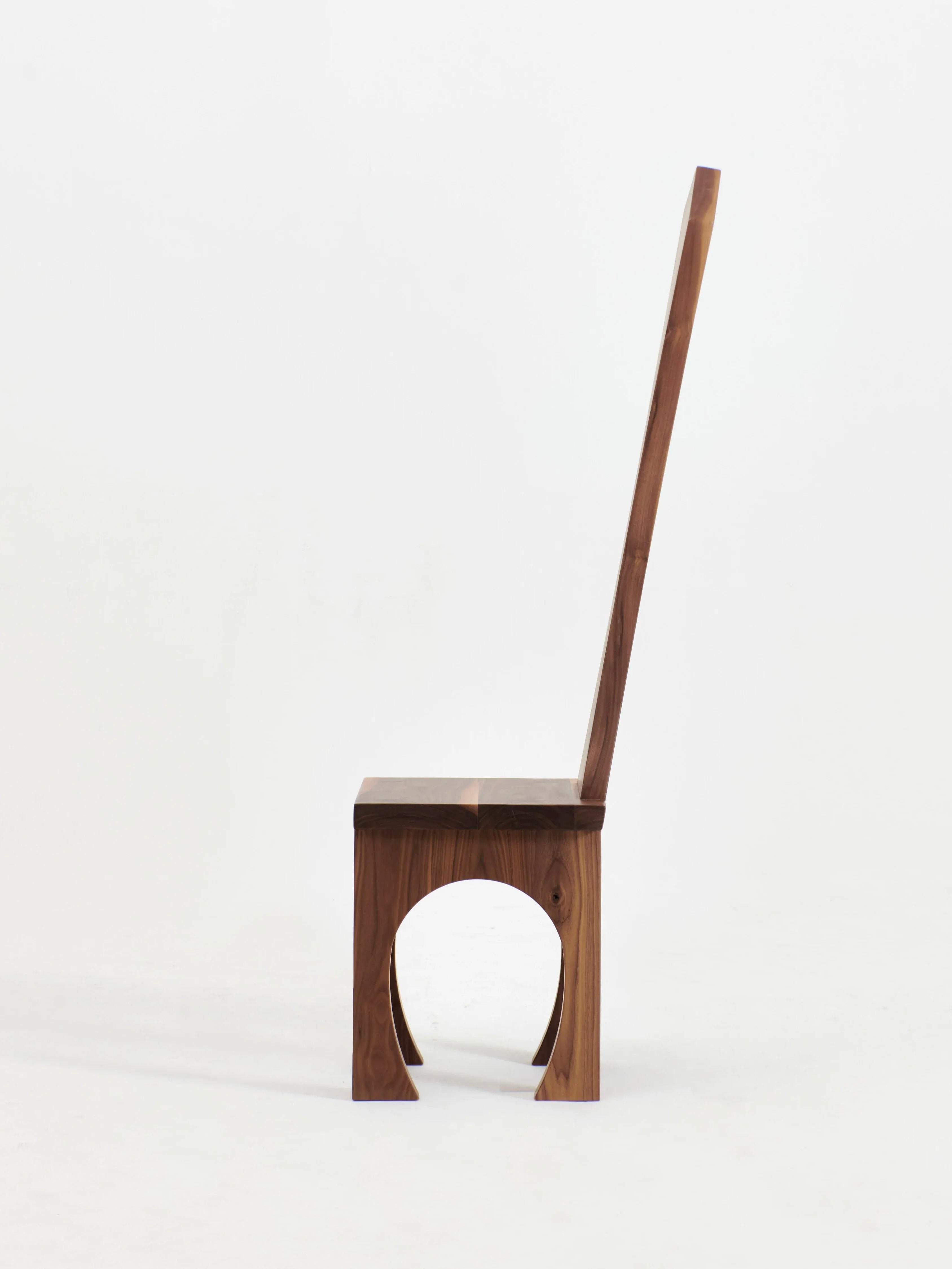 A side view of the Portugal Chair No 8 by Project 213A reveals its hand-carved wooden structure, characterized by a tall, straight backrest. This design highlights a unique circular cutout in the lower part of the seat, creating a minimalist and modern look. Crafted from solid walnut, the wood showcases a natural, polished finish against a plain white background.