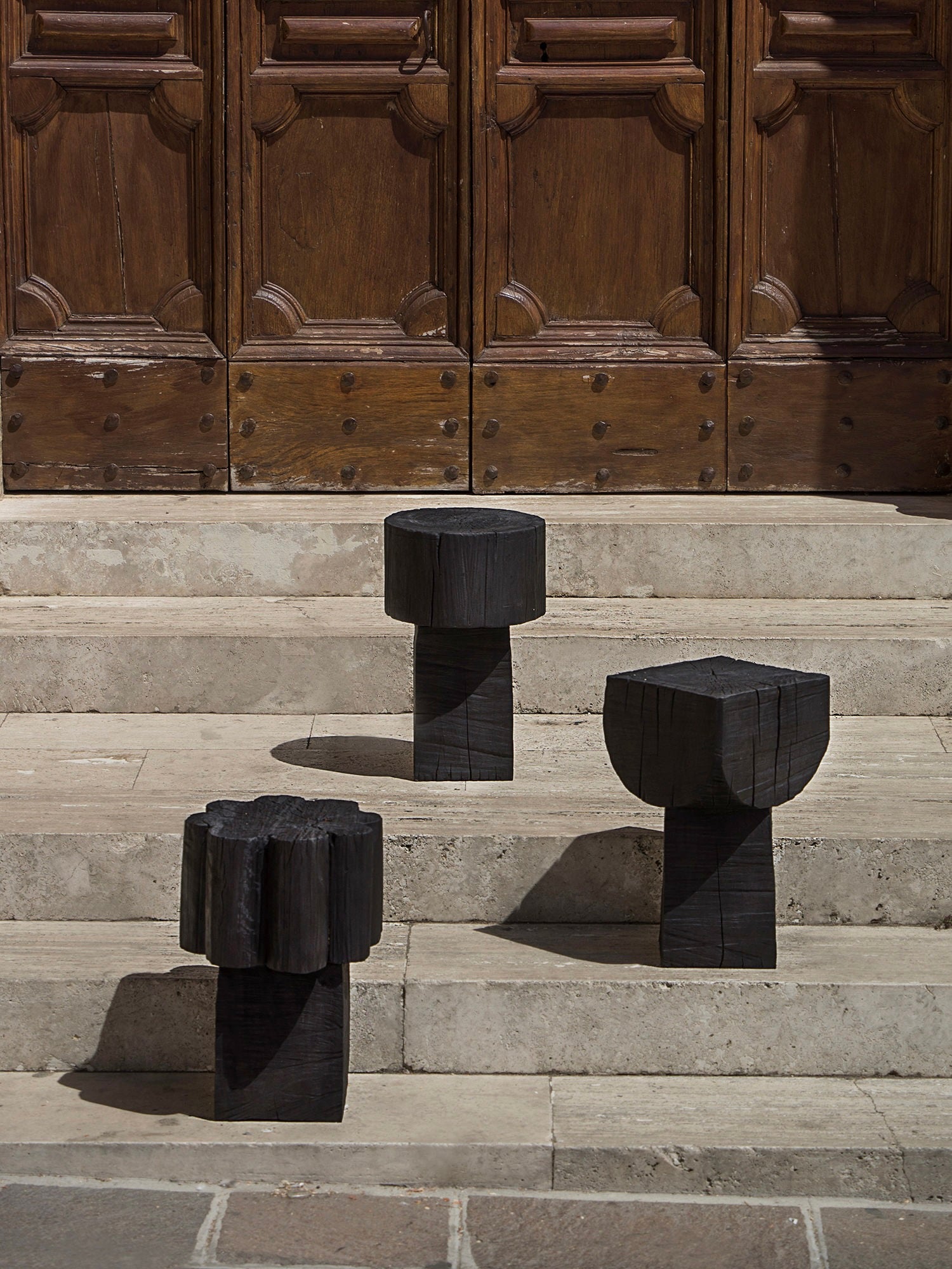 Three Burnt Black Coffee Tables/Stools by Touch With Eyes are positioned on stone steps in front of a large, ornate wooden door with decorative panels. Their unique shapes and textures complement the smooth stone and intricately carved wood backdrop.