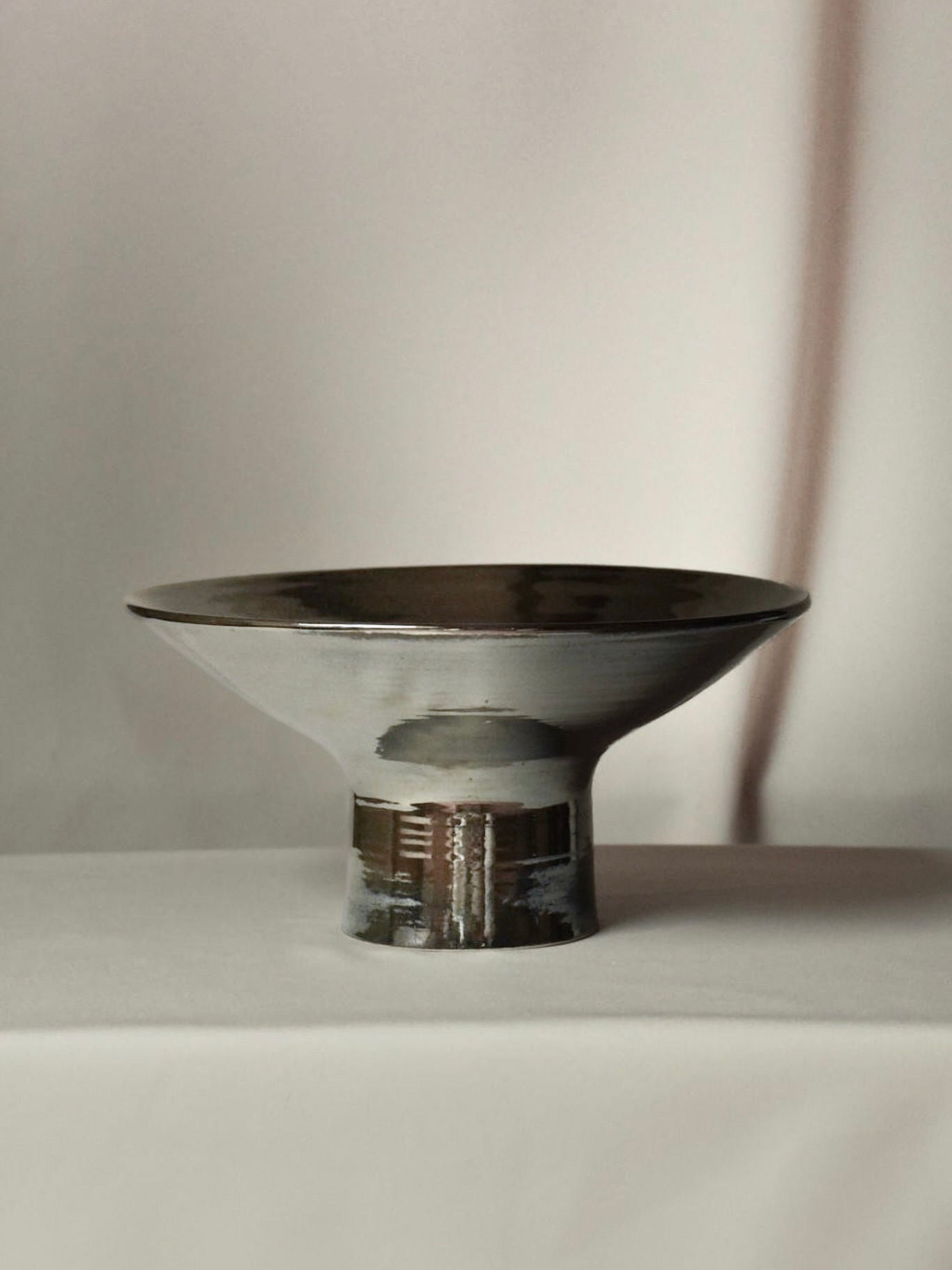 The Pedestal Centerpiece by Landy Rakoto Ceramics, featuring a metallic chrome finish in a modern style with a wide rim and narrow base, elegantly serves as a footed centerpiece on a neutral surface against a softly lit beige background.