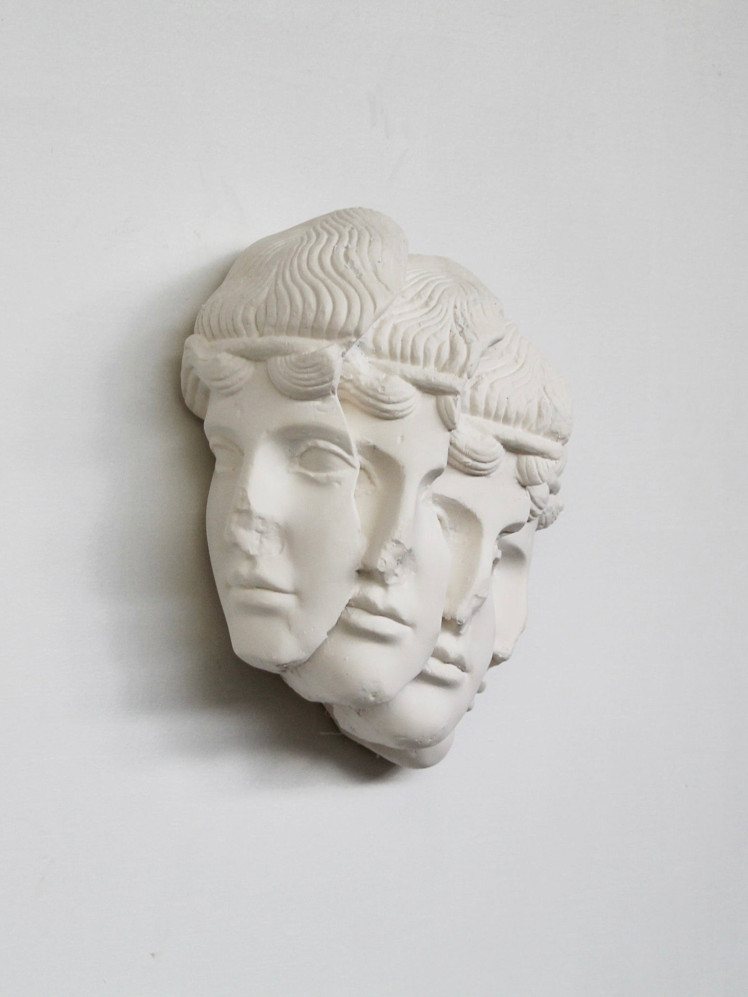 The "Empilement Mural" Sculpture by Marina Mankarios beautifully features a fragmented classical head with three overlapping faces, each with unique expressions. Made from white plaster art, it is mounted on a flat background that casts soft shadows, enhancing its three-dimensional charm. This piece is made to order for bespoke elegance.