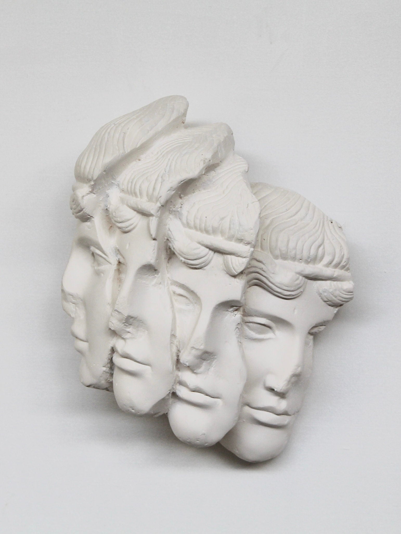 The "Empilement Mural" Sculpture by Marina Mankarios is a custom-made art piece in white plaster that showcases four overlapping male faces with classical features. Arranged in sequence, it produces an optical illusion against a simple white background.