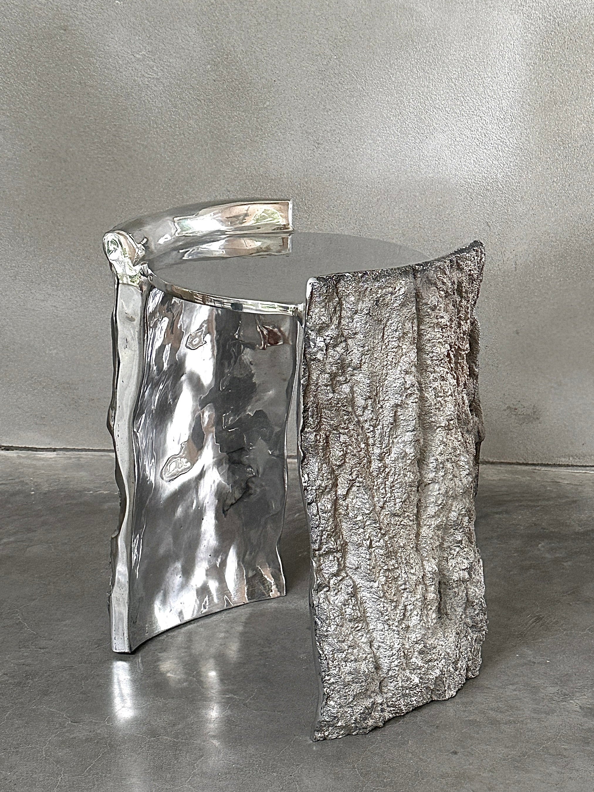 Kaytar's "Bark" Side Table features an Ironbark texture that mimics tree bark, with a high-gloss metallic finish on one side and a rough, bark-like surface on the other. This striking design contrasts beautifully when placed on a smooth, grey floor.