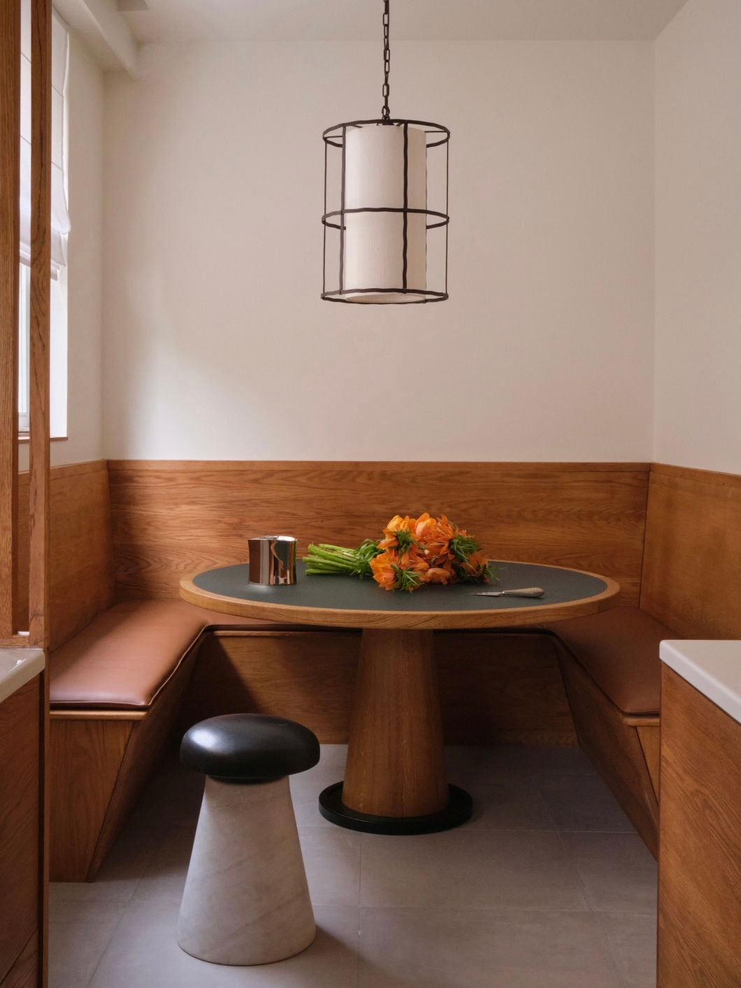 A cozy dining nook is adorned with the Jack Table by HAUVETTE & MADANI, showcasing its round wooden surface supported by a central leg. The table is surrounded by wooden benches topped with brown cushions. A contemporary black and white pendant light hangs overhead, casting light on a vibrant bouquet of orange flowers and a small cup placed on the table.