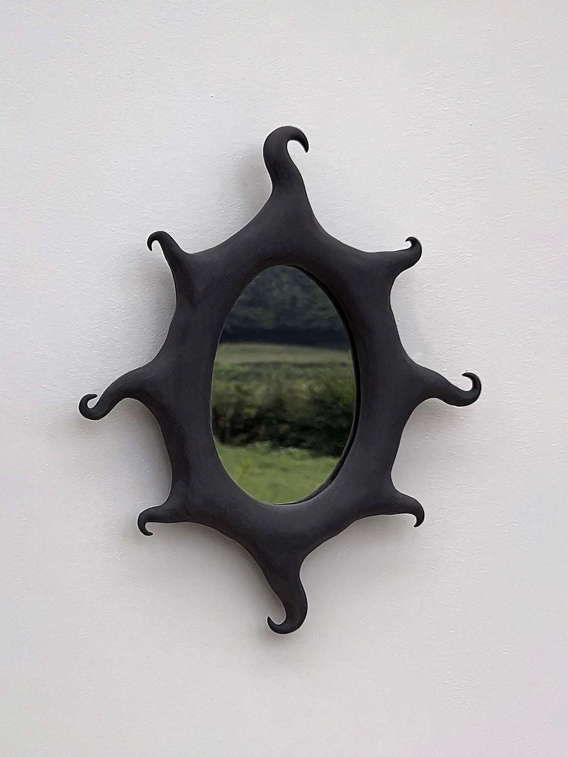 A decorative piece, the Black Sun Mirror by Solenne Belloir, featuring an irregular, wavy black matte finish frame, is displayed on a white wall. It reflects a blurred vision of greenery and trees outside, adding a magical presence to the room.