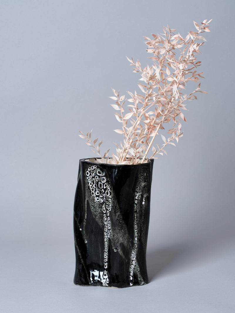 Against a plain background, the Swirly Vase Black and White by Astrid Öhman, handmade and glossy with a wavy surface, holds pale dried branches with delicate leaves, contrasting colors and textures.