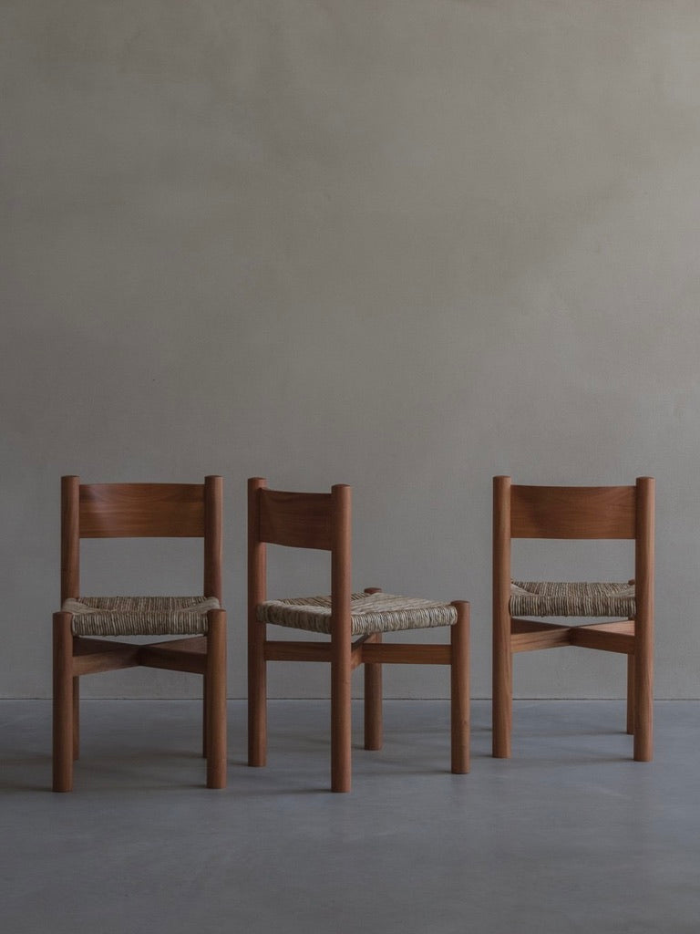 Three 'Les Allues' straw chairs by Bicci de' Medici, featuring exquisite craftsmanship with woven seats, are arranged in a row on a smooth concrete floor. The background showcases a plain light gray wall, creating a minimalist and modern aesthetic.