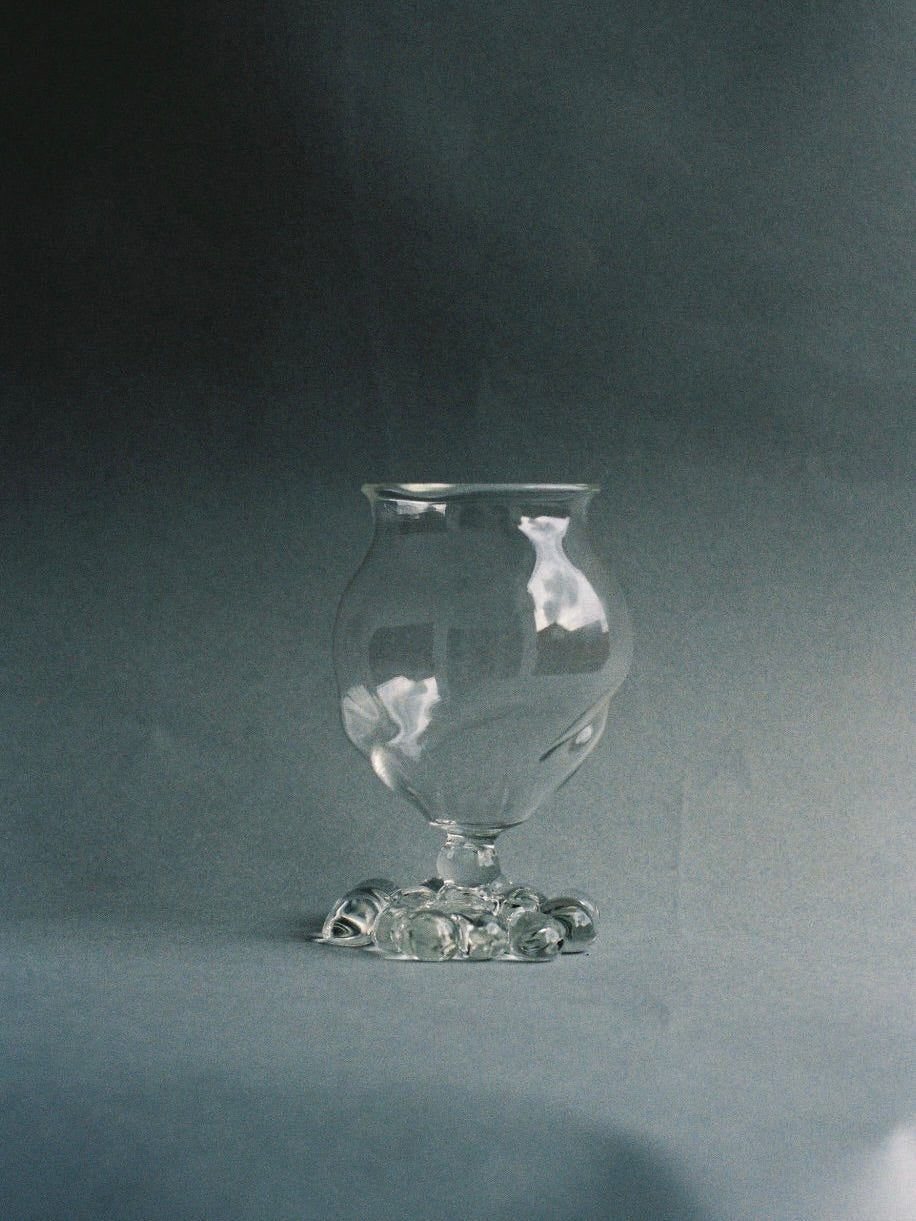 A clear, intricately designed water glass goblet from the Justine Menard collection stands against a soft gray background. The hand-blown goblet has a rounded bowl with unique bubble details and an ornate, decorative base. The image highlights the elegance and detailed craftsmanship of these Mouth Blown Water Glasses - Set Of Two.