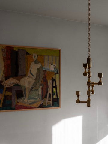 A painting of an abstract figure is displayed beside a large Sculptural Brass Chandelier by Spigel on a plain wall. Sunlight casts a shadow across the wall, highlighting both the artwork and the fixture.