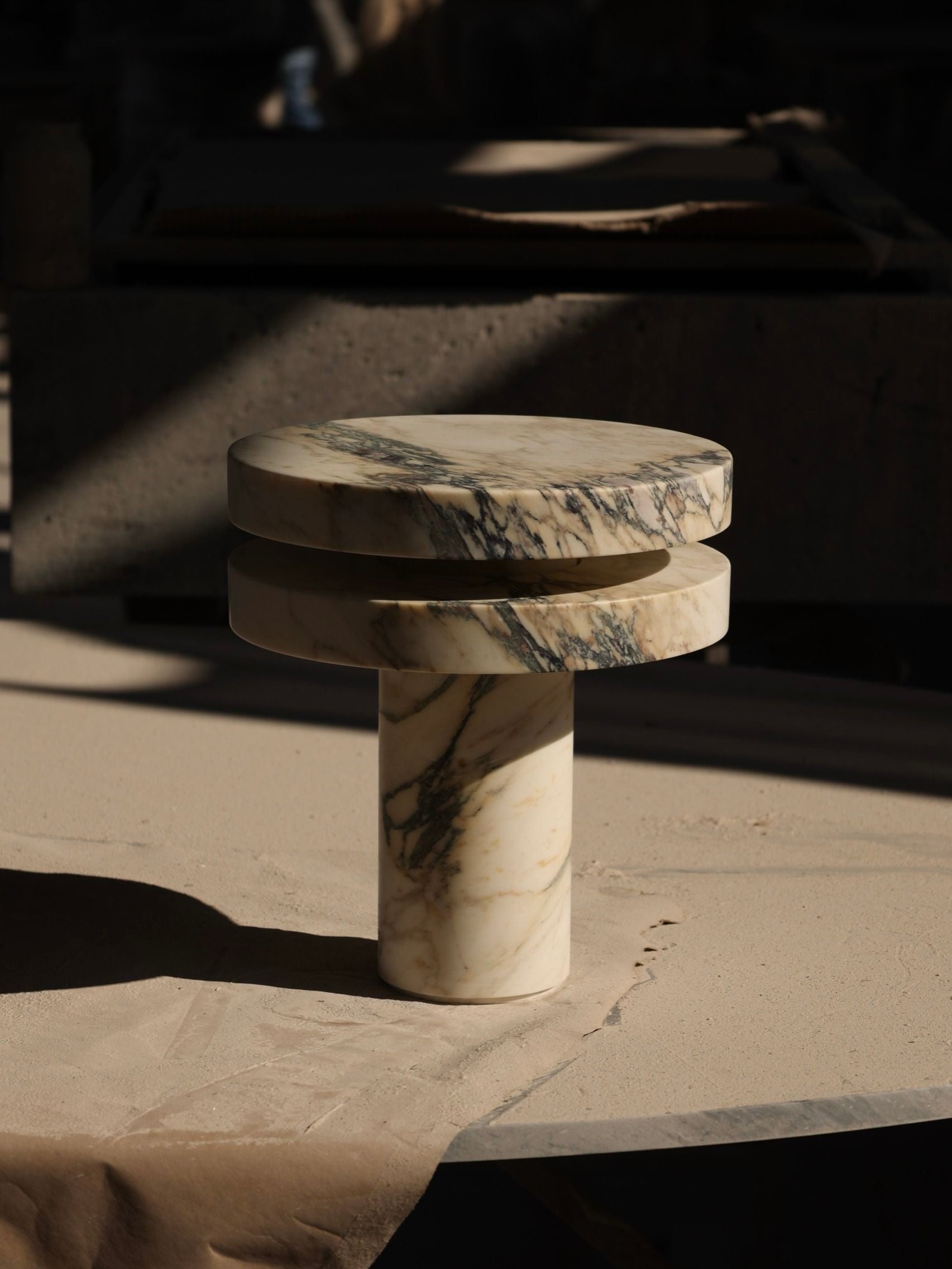 The 'M_014' Marble Table Lamp by Monolith, featuring a small round pedestal with two stacked disk layers on a cylindrical base, rests on a sandy surface. Its white and gray veining hints at brutalist design, with soft shadows suggesting sunlight filtering from above.