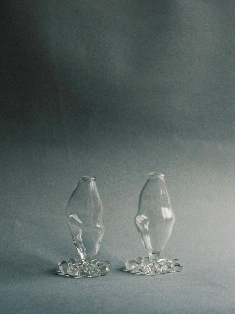 Two abstract, shapeless vases from Justine Menard stand on a grey backdrop. Each mouth-blown Small Vase sits atop a small base made of clear, irregularly shaped glass pieces. This modern and minimalist piece seamlessly blends into any home decor.