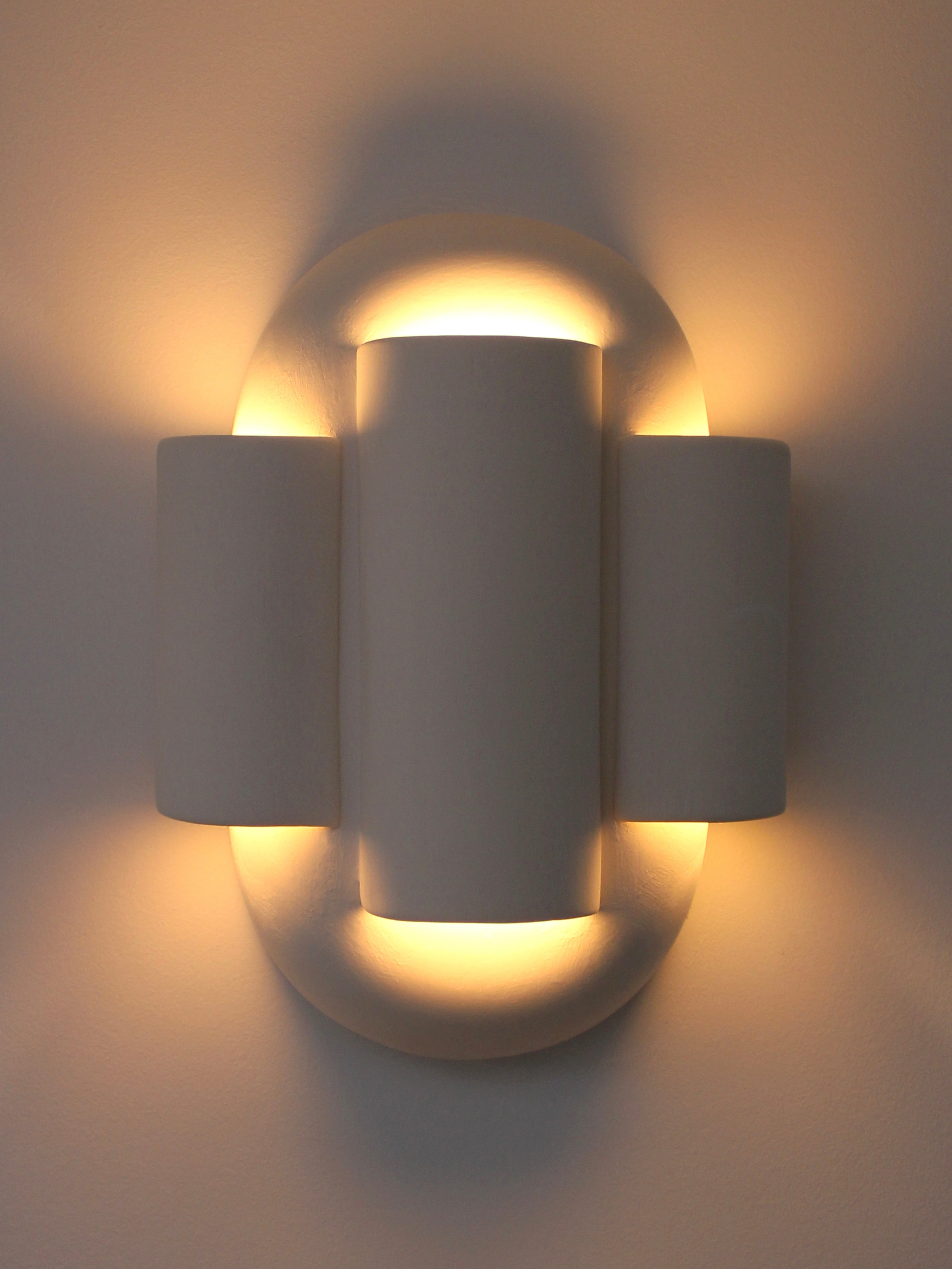 The YEREK Wall Light from Villa Arev features a contemporary design with a cylindrical center and two symmetrical extensions on either side, all in a sleek, matte white finish. The light gently diffuses around the fixture, casting warm, soft illumination against the wall.