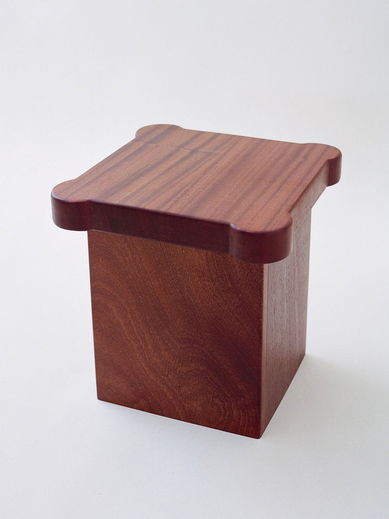 The Bijou Mini Table by MUHLY is a wooden, cube-shaped box with a flat, slightly decorative lid. Made from polished wood, the box showcases its natural grain and rich brown color. Perfect as a bijou mini table or plant stand, the lid features rounded corners that extend slightly beyond the edges of the box.