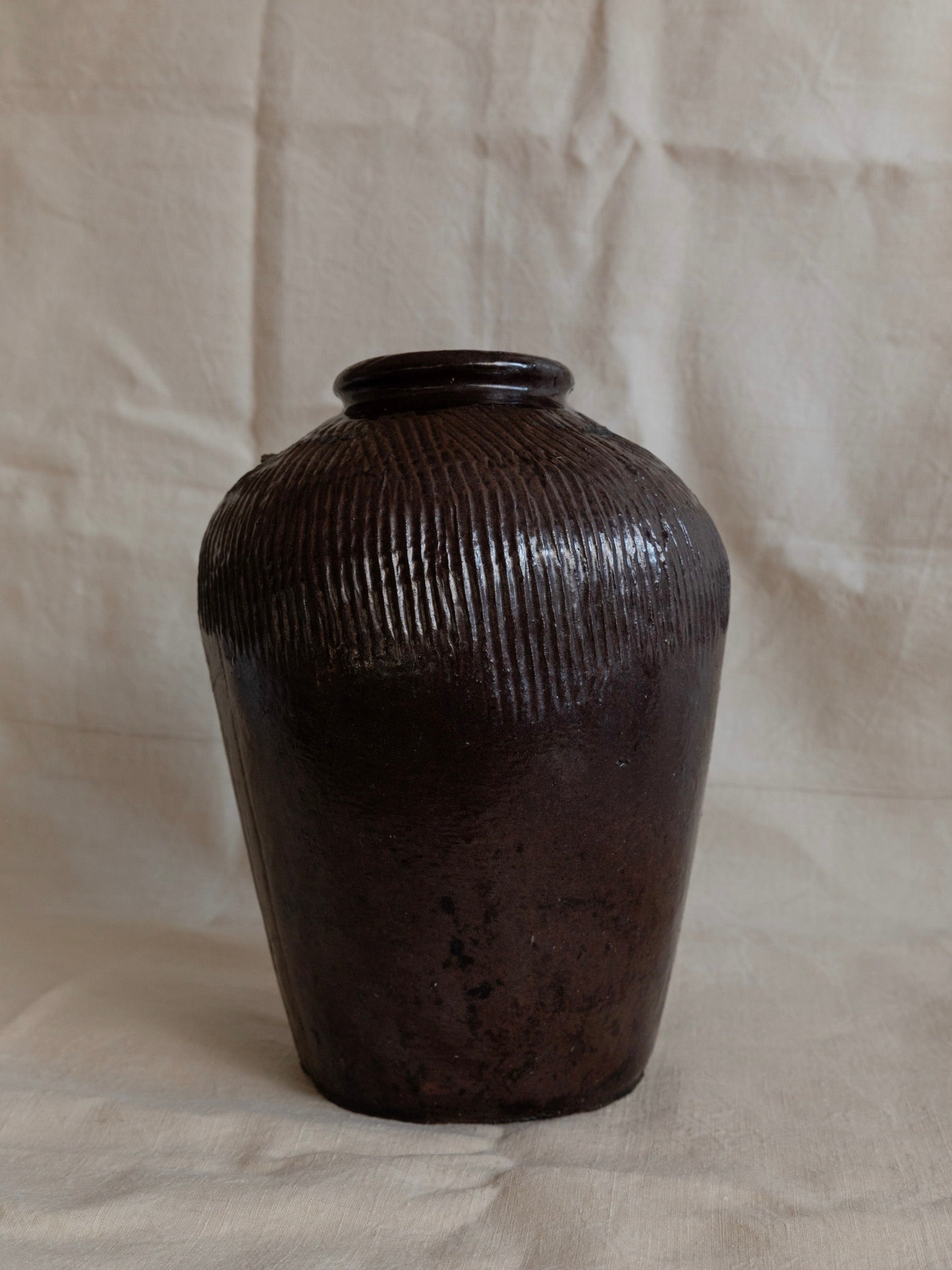 An Antique Chinese 19th Century Ceramic by Bottega Jacobs, showcasing a dark rustic brown hue with a narrow neck and wide body, rests against a plain, light-colored fabric background. Its ribbed design evokes the elegance of an antique Chinese rice wine pot, exuding an earthy and rustic charm.