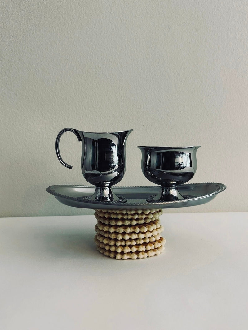 A polished Vintage Alessi Sugar & Creamer Set by Flock Studio sits gracefully on a coordinating tray, perfectly complementing each other. The tray is beautifully balanced on a stack of wooden beads against a clean white backdrop.