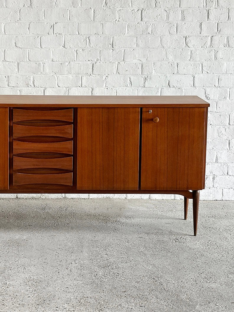 The Introverso Italian Modernist Teak Sideboard, crafted in 1960s in the style of Vittorio Dassi, features a natural finish with four drawers, two cabinet doors with round handles, and stands on tapered legs against a white brick wall and concrete floor.