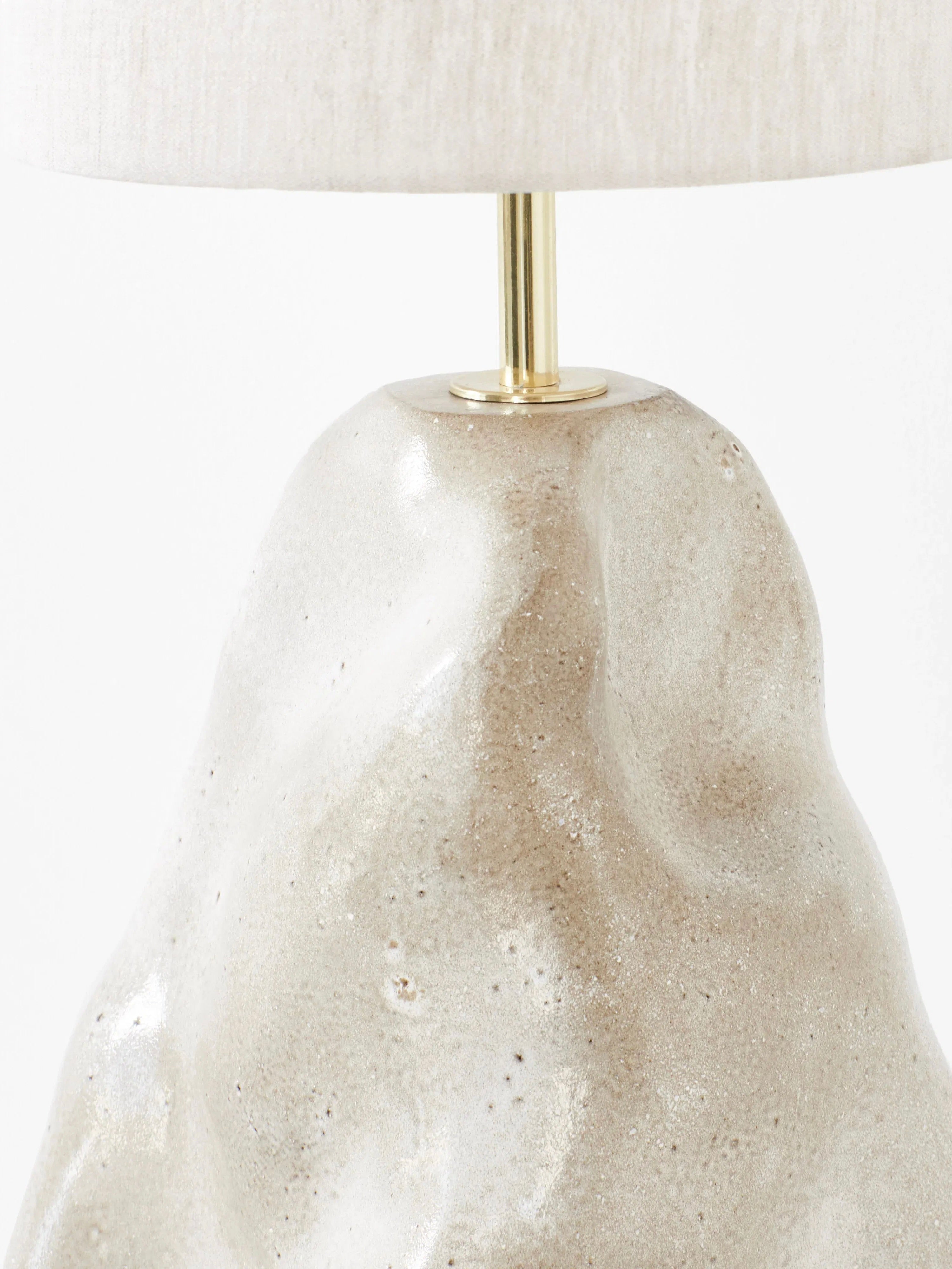 A close-up of the "Menina" Floor Lamp in White by Project 213A, featuring a textured, irregularly shaped beige ceramic base and a simple, cylindrical beige fabric lampshade. The lamp showcases traditional craftsmanship with a minimalistic design and a subtle metallic accent at the connection point between the base and the shade.