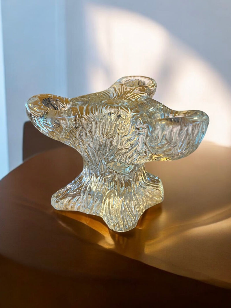 The Sculpted Glass Candelabra by Flock Studio, featuring an abstract, organic design, sits on a smooth light brown surface. It captures warm light and casts intricate shadows.