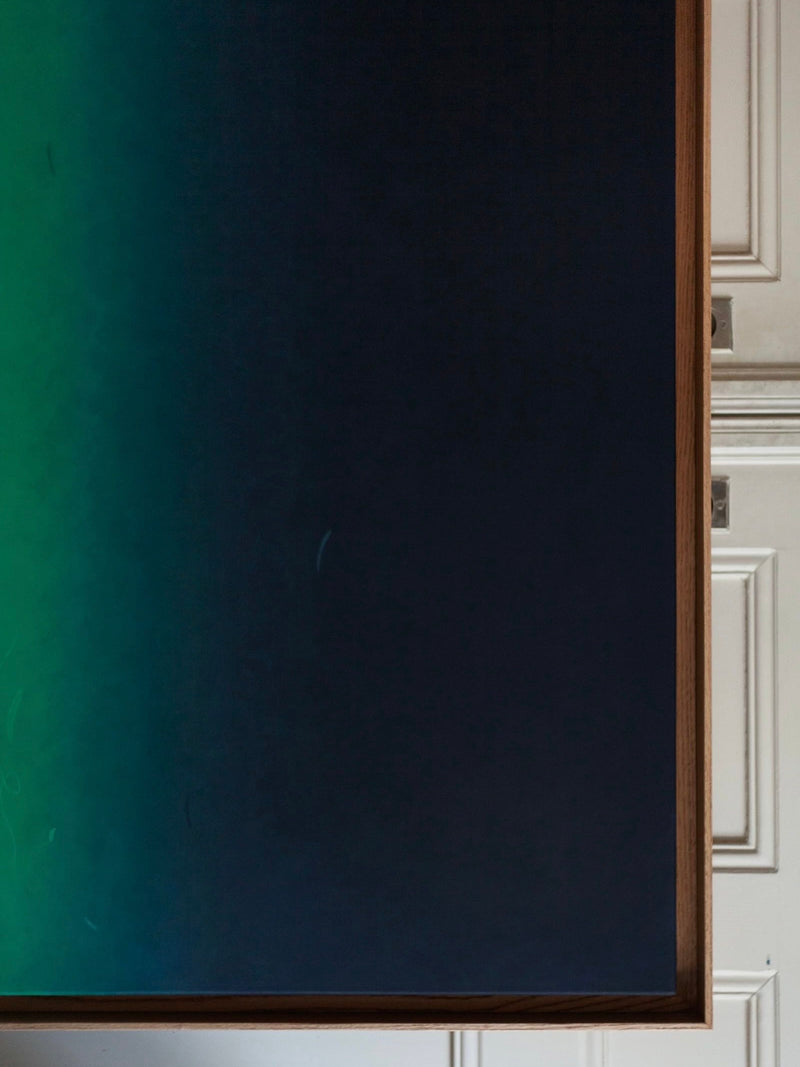 The art piece "Beneath and Below, 2024 Acrylic on Canvas" by Rebecca von Matérn is showcased on a wall, displaying a gradient that shifts from dark green on the left to dark blue on the right. This minimalist artwork is framed with a shadow frame made of hard waxed oak, set against a white paneled background.