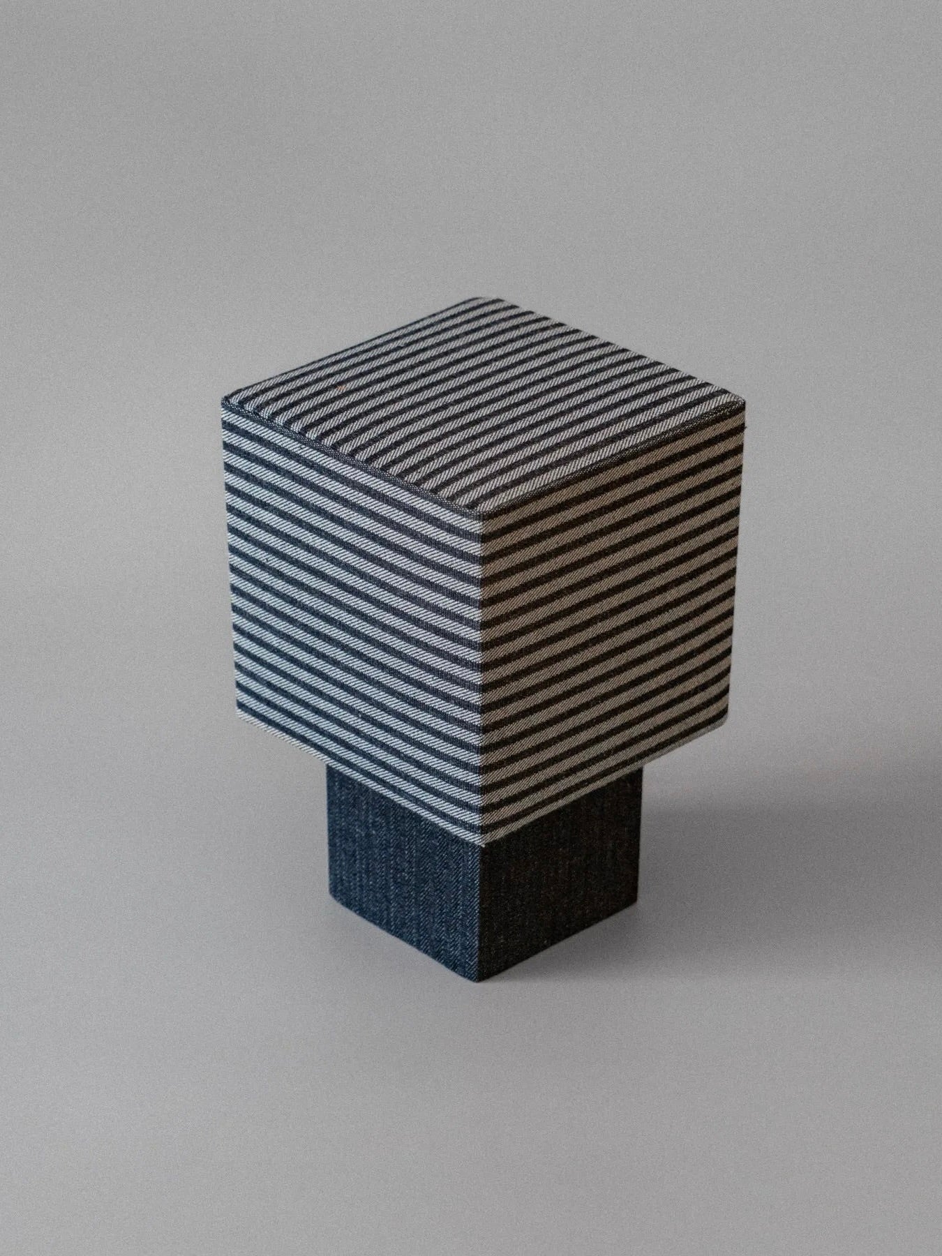 A contemporary, cube-shaped table lamp with a striped denim shade crafted from deadstock fabrics and a solid dark base by mattina moderna is displayed against a neutral gray background.