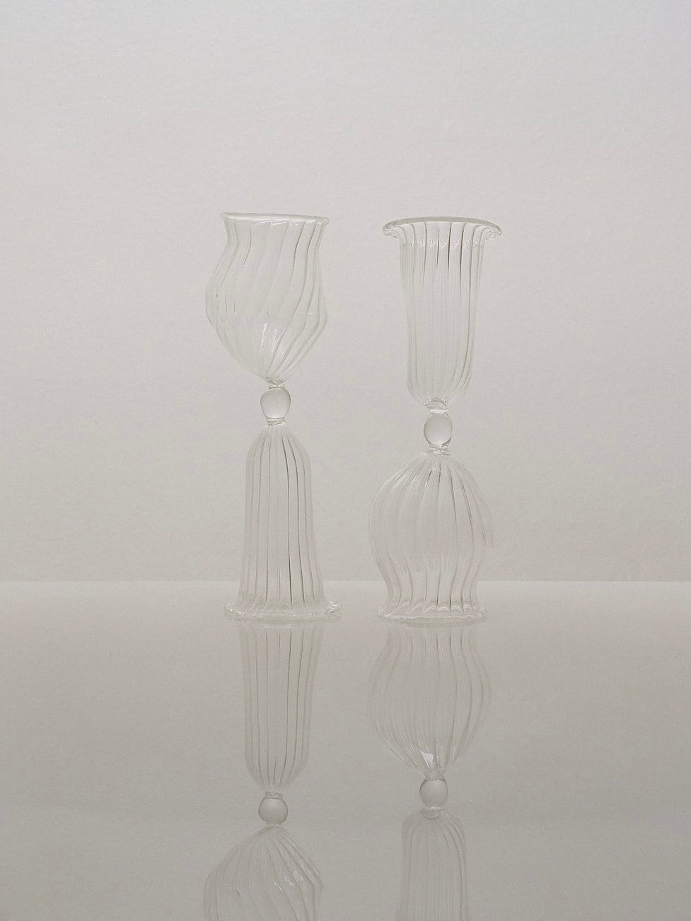 The Cléodore Glass set of two, crafted by Justine Menard, features intricately designed, clear mouth-blown glass goblets with elongated stems and a vertical ribbed pattern. They stand side by side on a reflective surface against a neutral background, highlighting the delicate craftsmanship and classic design of these exquisite home decor pieces.