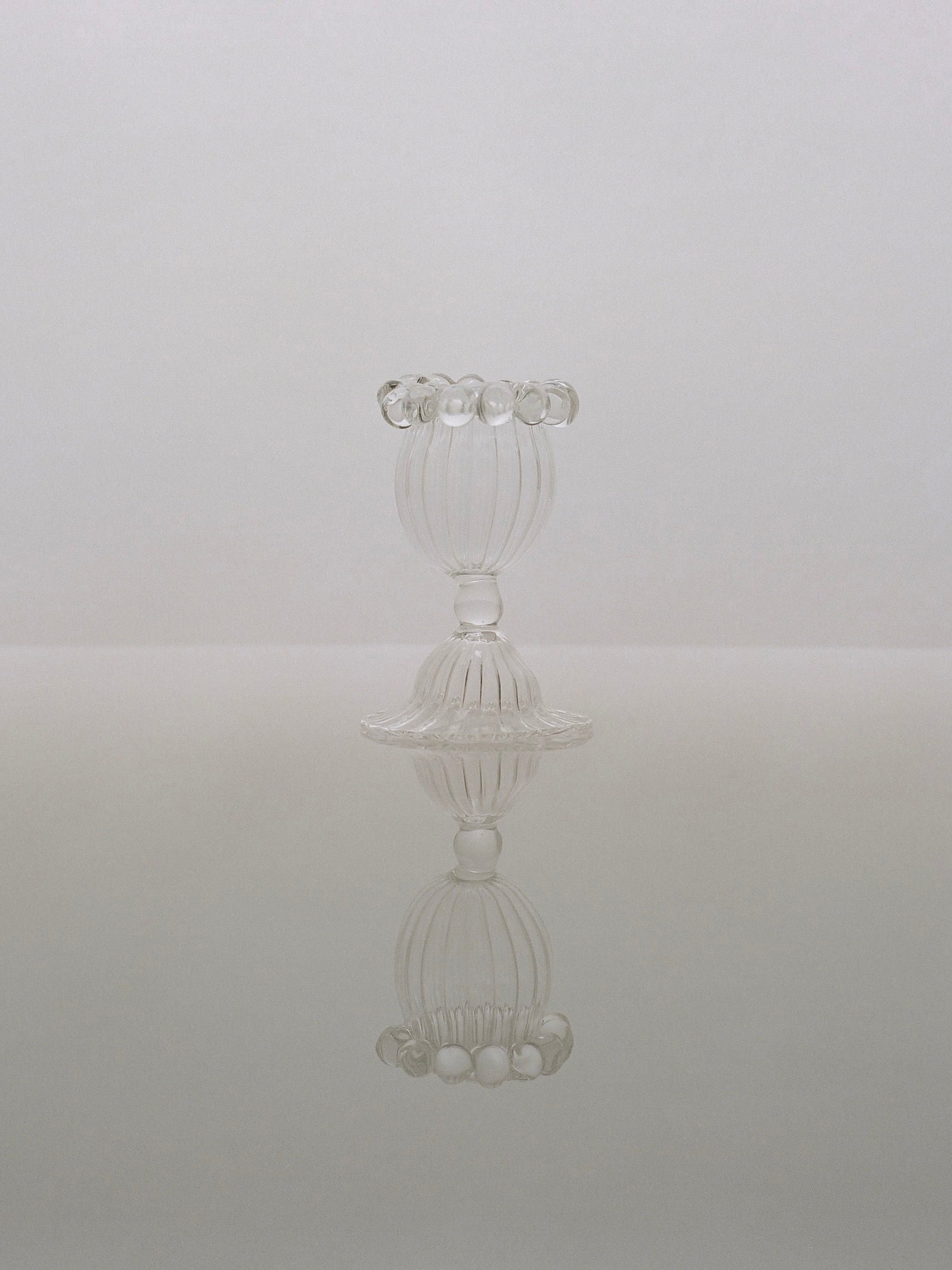 A pair of Eudore Egg Cups by Justine Menard is centered against a light, gradient background. The clear, intricately-designed egg cups' reflections are visible on the glossy surface beneath them, creating a symmetrical, mirror-like image reminiscent of hand-blown goblets' artistry.