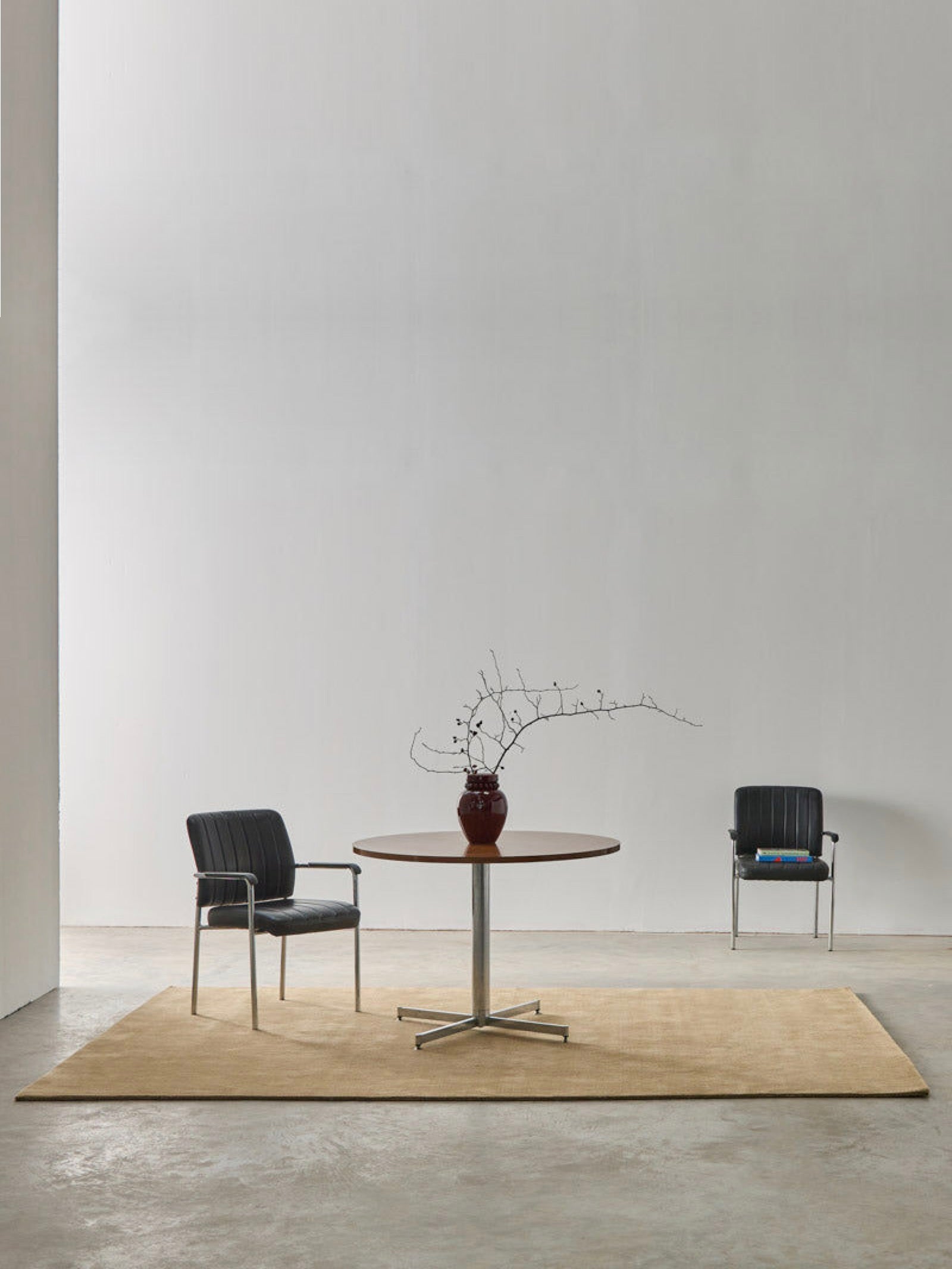 A minimalist room with two black chairs, a round wooden table with metal legs, and MOST's 'Châtaigne' hand-woven rug in New Zealand wool. A vase of bare branches sits on the table against a plain white wall.