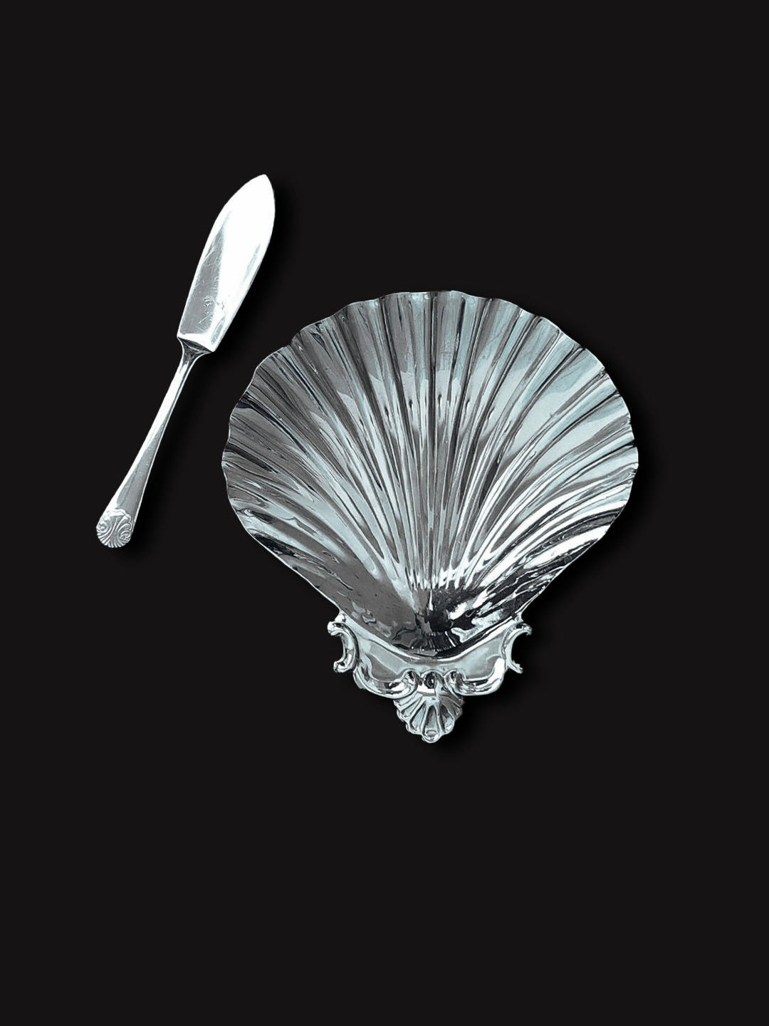 The Shell Butter Dish by Les Objoies, featuring a silver shell-shaped design with a detailed edge, is complemented by an assorted stainless serving knife. Their intricate designs stand out against the black background.