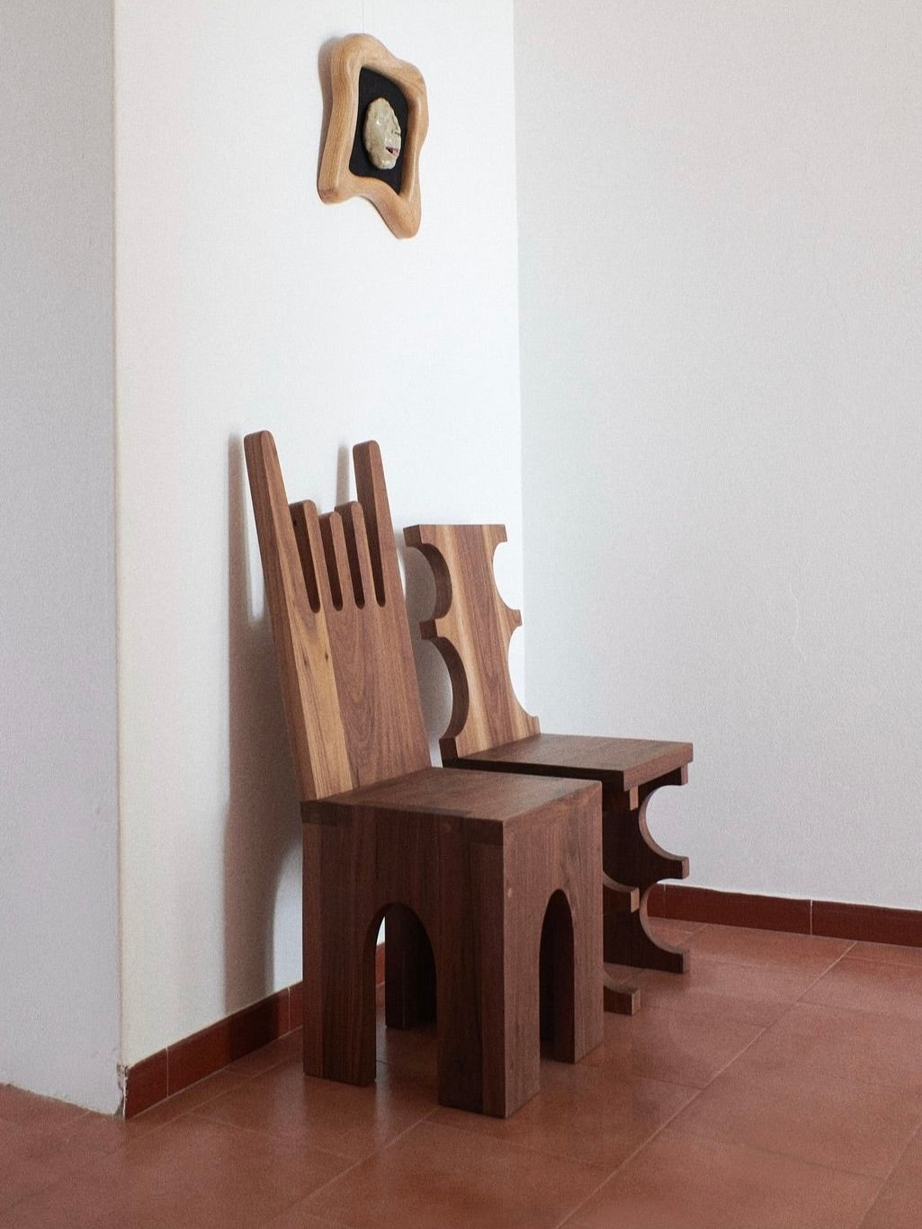 Two unique wooden chairs, including a Portugal Chair No 5 by Project 213A crafted from solid walnut, stand against a plain white wall on a tiled floor. One chair has a tall, irregularly-shaped backrest, while the other boasts graphic cutout shapes in its backrest and sides. A carved face hangs above.