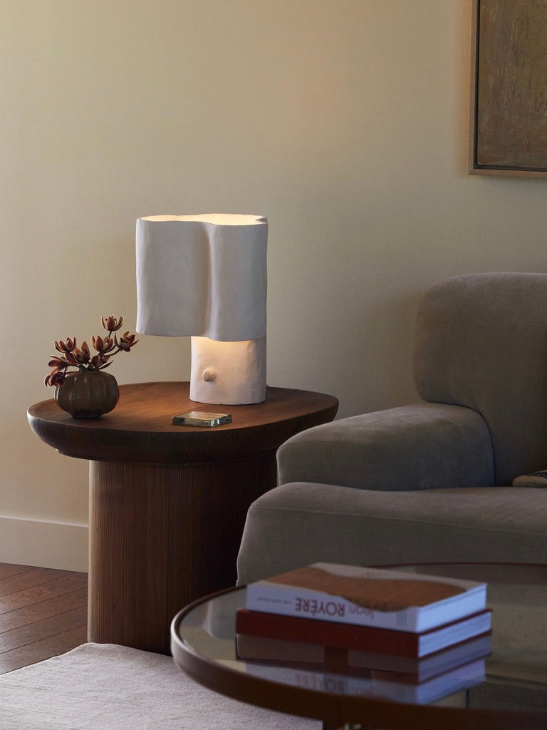 A cozy living room showcases a modern beige couch, a round wooden side table with a handcrafted Studio Tristan Kallas Cantilever Freeform Table Lamp, plus a small plant and smartphone. A round glass coffee table displays two stacked books, while light-colored walls enhance the warm ambiance beautifully.