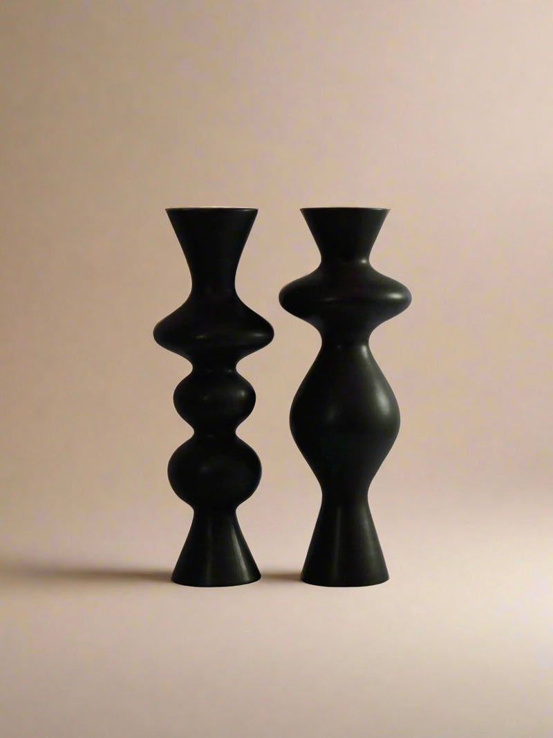 Against a neutral background, the Pair of Ceramic Candleholders by Ransom & Dunn stands side by side, their black, abstract forms bringing to mind the female silhouette. The candleholder on the left showcases a series of round bulges, while its counterpart on the right boasts a more curvaceous form reminiscent of elegant vases.