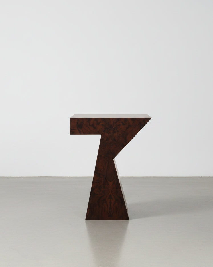 The "Columbina" Side Table in Walnut Burl Veneer by Animate Objects features a unique Z-shaped design and dark wood finish, handcrafted in Istanbul, shown on a polished brown floor against a plain white wall.