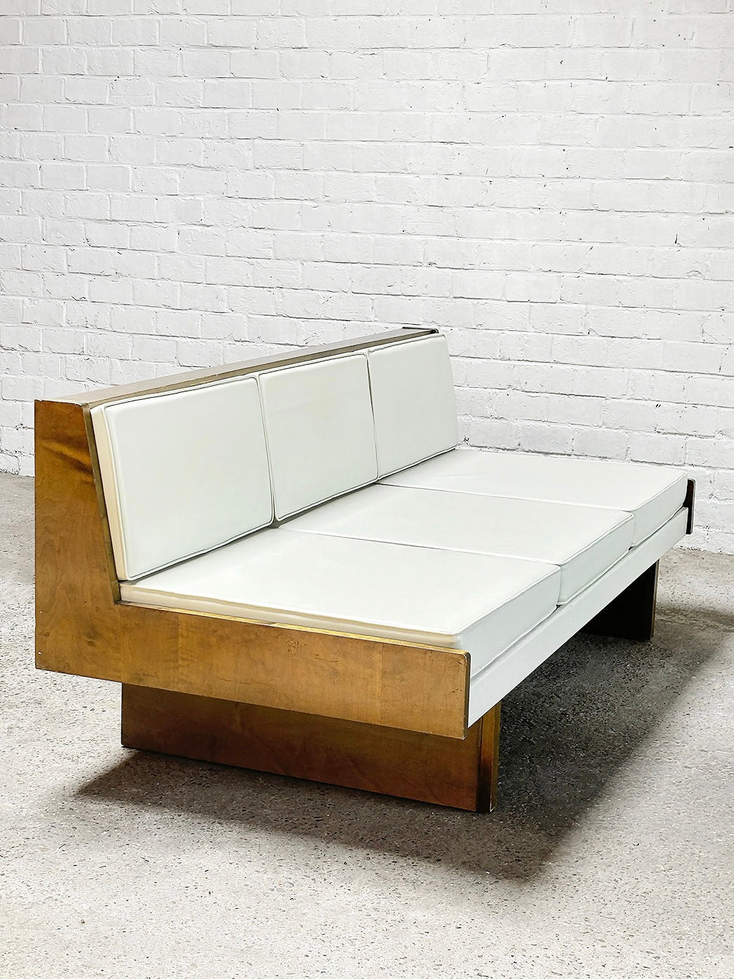 The French Modernist Sofa Set in White Leather by Introverso, a minimalist design from the 1950s, features three back cushions and a spacious seat with a wooden base against a white brick wall. The concrete floor adds an industrial vibe for an elegant contrast.