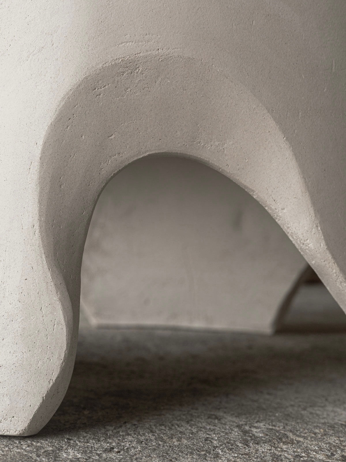 Close-up of the Lac - Carved Table by Lea Bigot in sandstone and oak, featuring an abstract off-white sculpture with a smooth, curving surface and arched opening atop a textured speckled gray base, exemplifying modern design with a shadow enhancing its elegance.