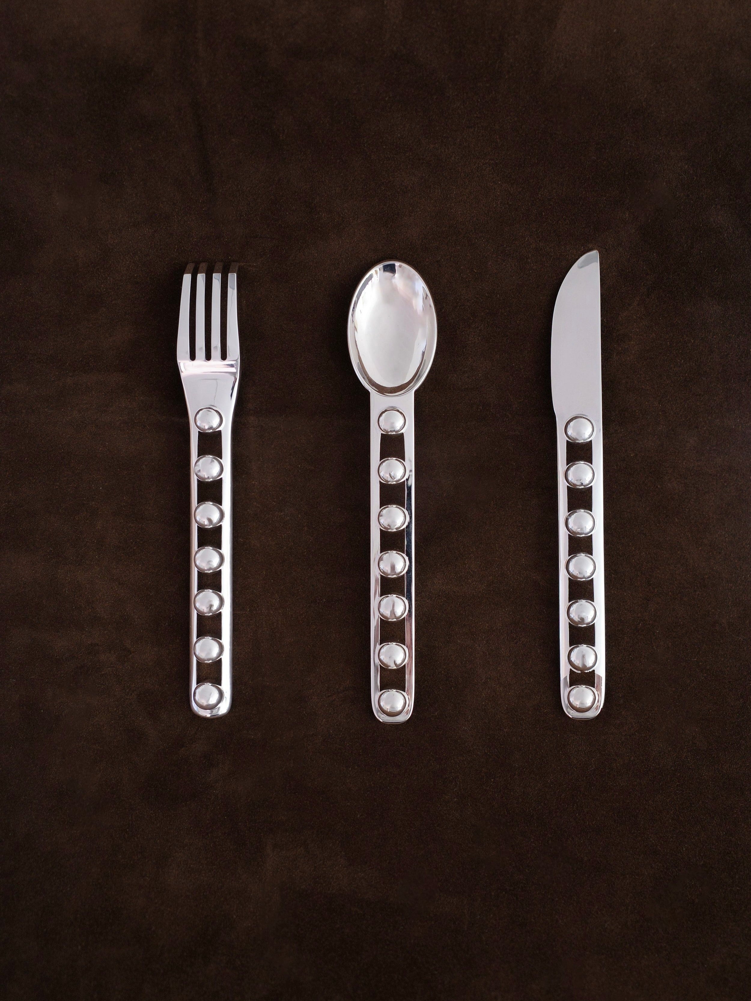 The "Seven Ball" Cutlery Set by Jessi Burch lies on a dark brown surface, showcasing a fork, spoon, and knife. The handles exhibit a design with circular cutouts, merging functionality with artistic elegance.