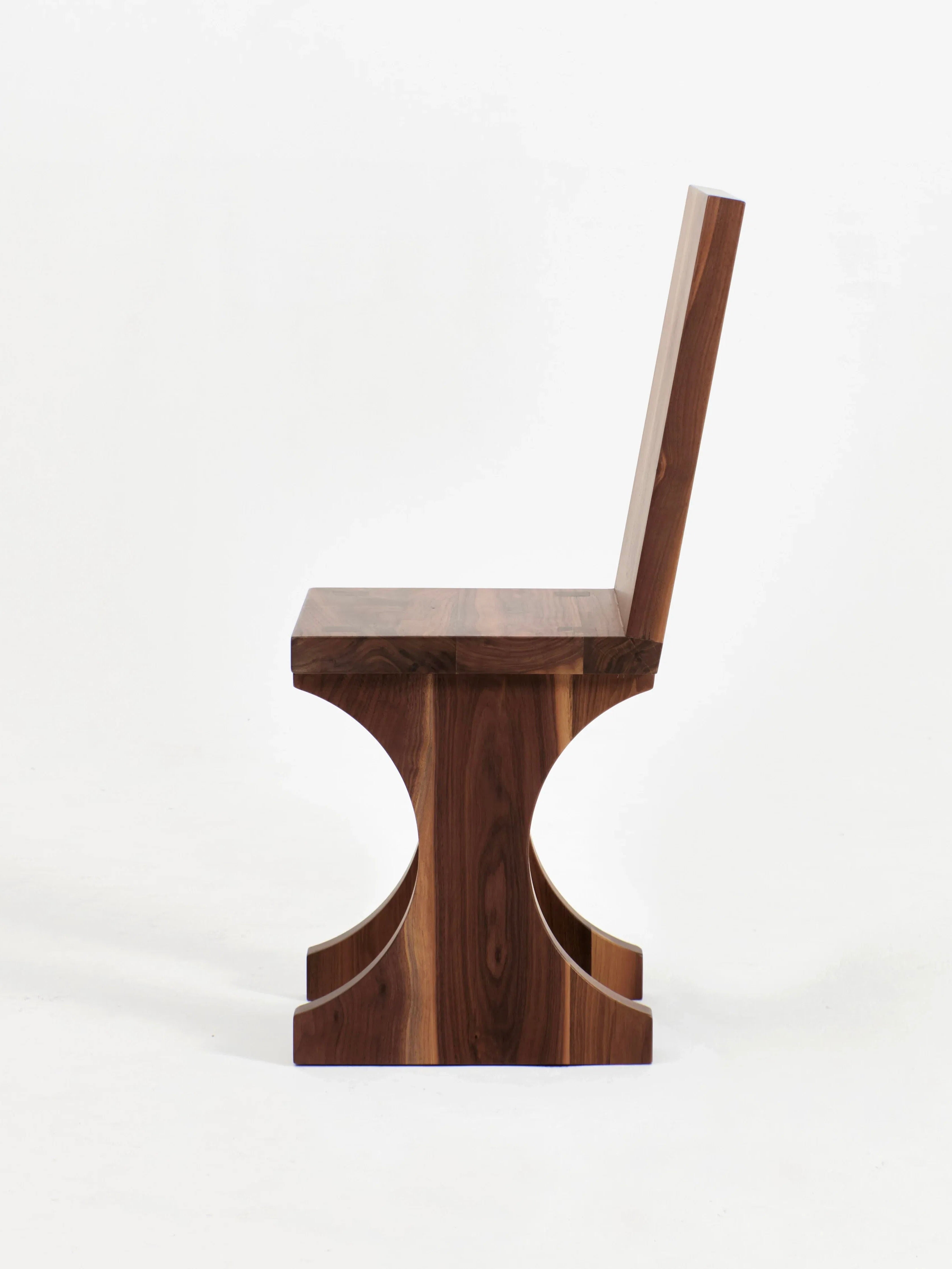 A side view of the Portugal Chair No 3 by Project 213A, a modern wooden chair featuring a minimalist design. This handcrafted piece showcases a straight backrest and seat with a distinctive base that has curved notches on both sides, offering a stylish, unique look. Made from solid walnut, the wood is smooth and polished to perfection.