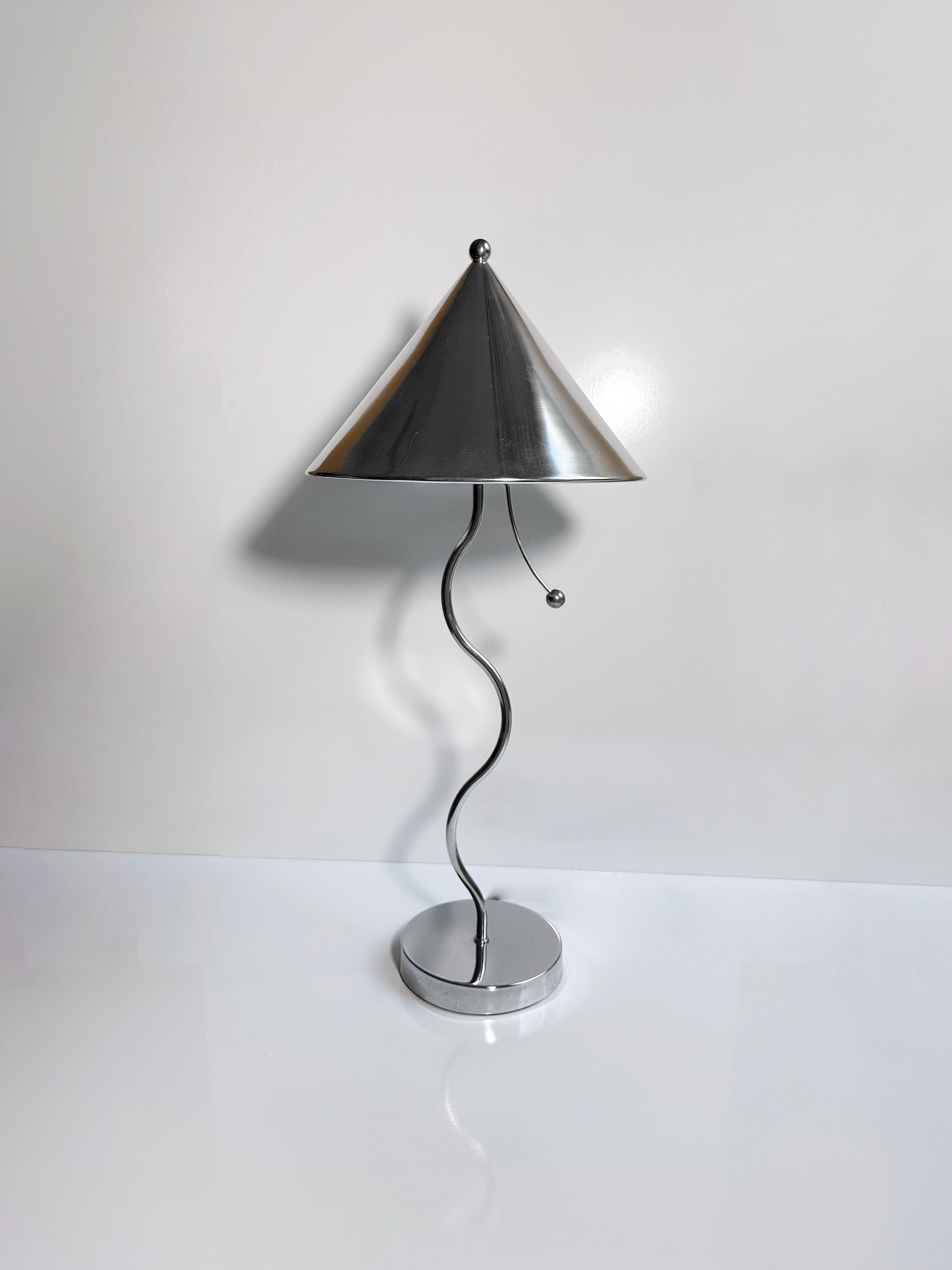 The Postmodern Wavey Chrome Table Lamp by Dodo Vintage features a sleek, wavy silver stem, a conical metallic shade, and chrome-plated metal finishes. This modern design includes a pull chain switch and a round base, beautifully showcased against a plain white background.