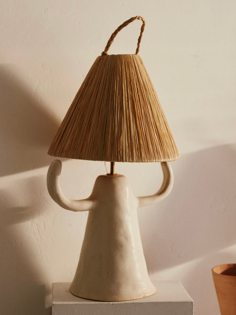 Introducing the Dona Table Lamp by marta bonilla, a distinctive piece with a stoneware base featuring two arm-like handles and a woven raffia shade. It beautifully casts soft shadows on light backgrounds, enhancing any room with an artistic flair.