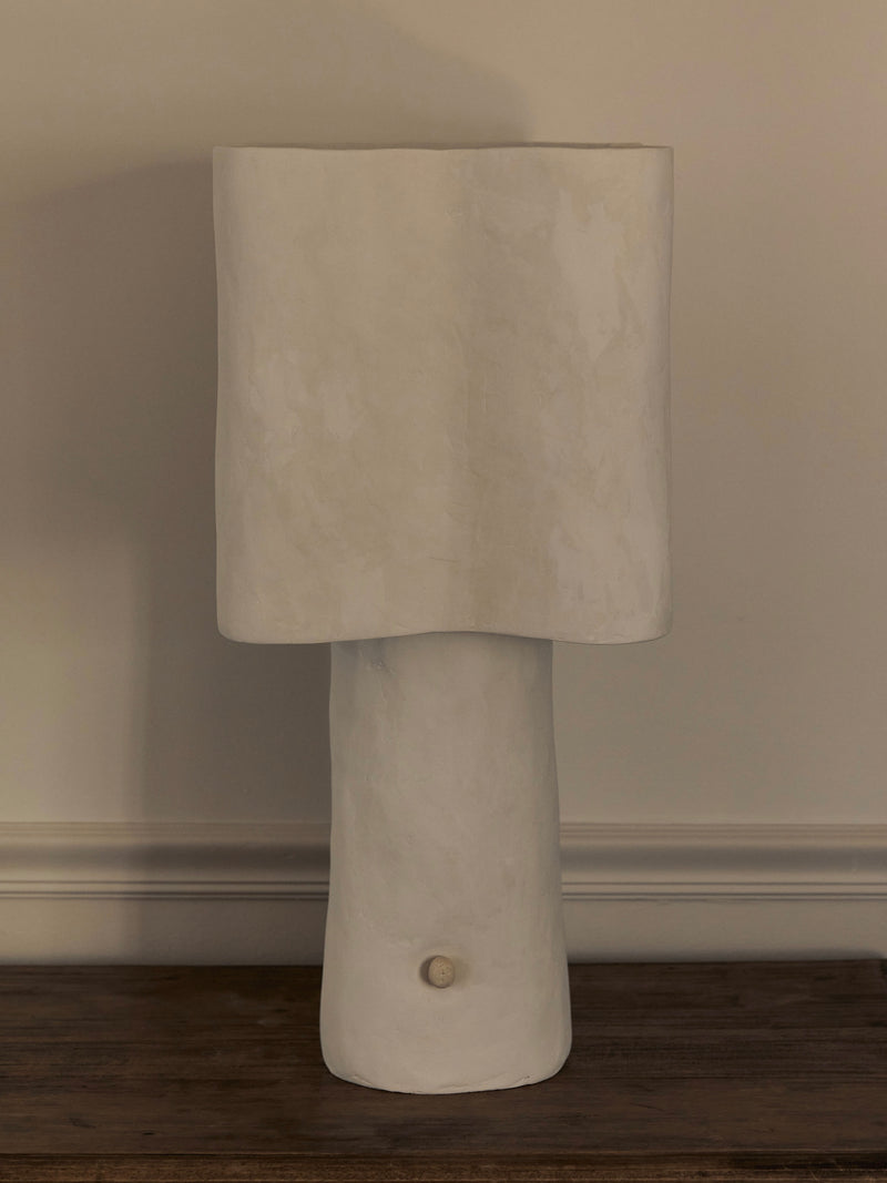 The Studio Tristan Kallas Freeform Table Lamp boasts a minimalist design with a smooth cylindrical base and tall rectangular shade in off-white, elegantly placed on a wooden surface against a plain wall.