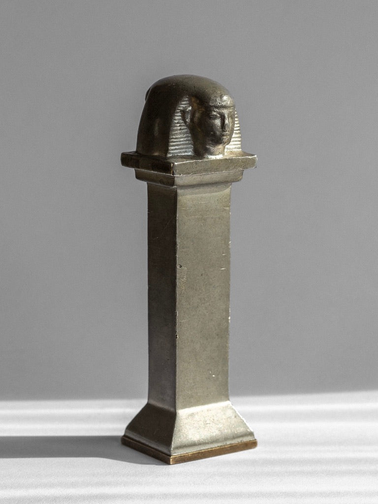 A tall, rectangular Art Deco Seal Stamp Sculpture from the 1930s, created in Sweden by Spigel, stands on a pedestal with a double-headed sphinx sculpted on top. The object appears to be made of pewter and brass with a smooth, weathered surface, set against a light grey background.