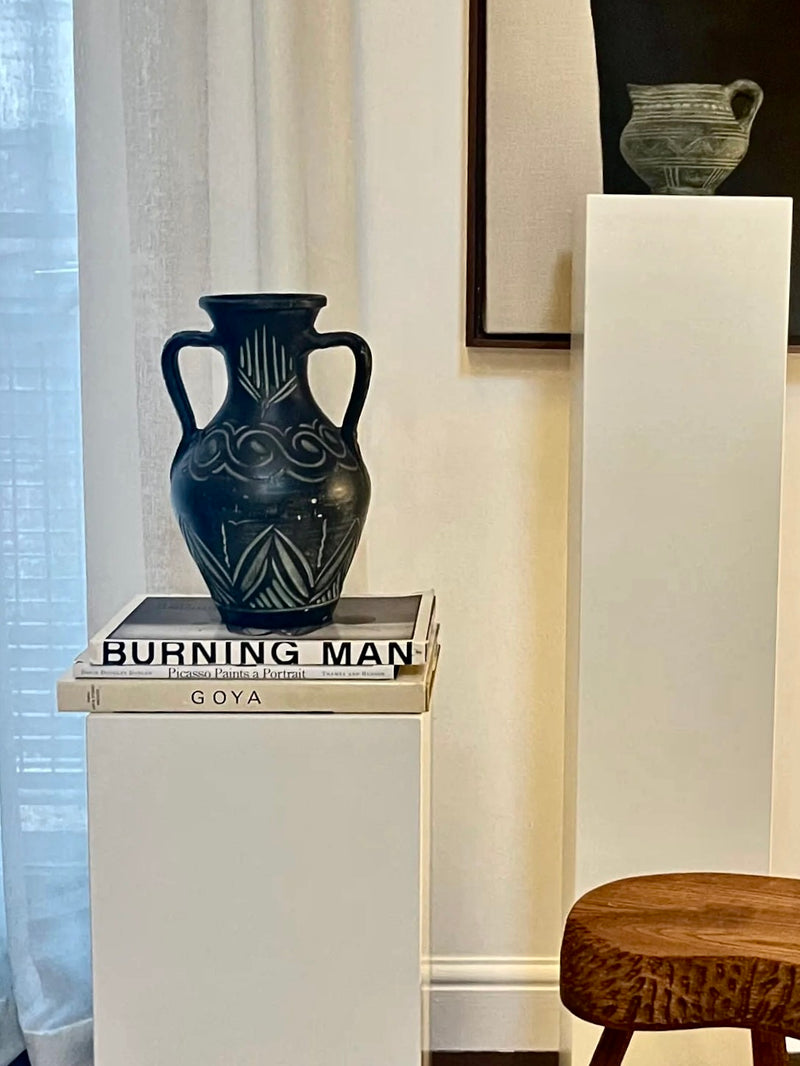 An elegant, unique European Large Carved Vase by Aesthete Label with two handles is placed atop a stack of two books titled "Burning Man" and "Goya." The books and vase are positioned on a white pedestal next to a window adorned with sheer curtains. Beside it, another smaller vase is showcased on a taller pedestal.