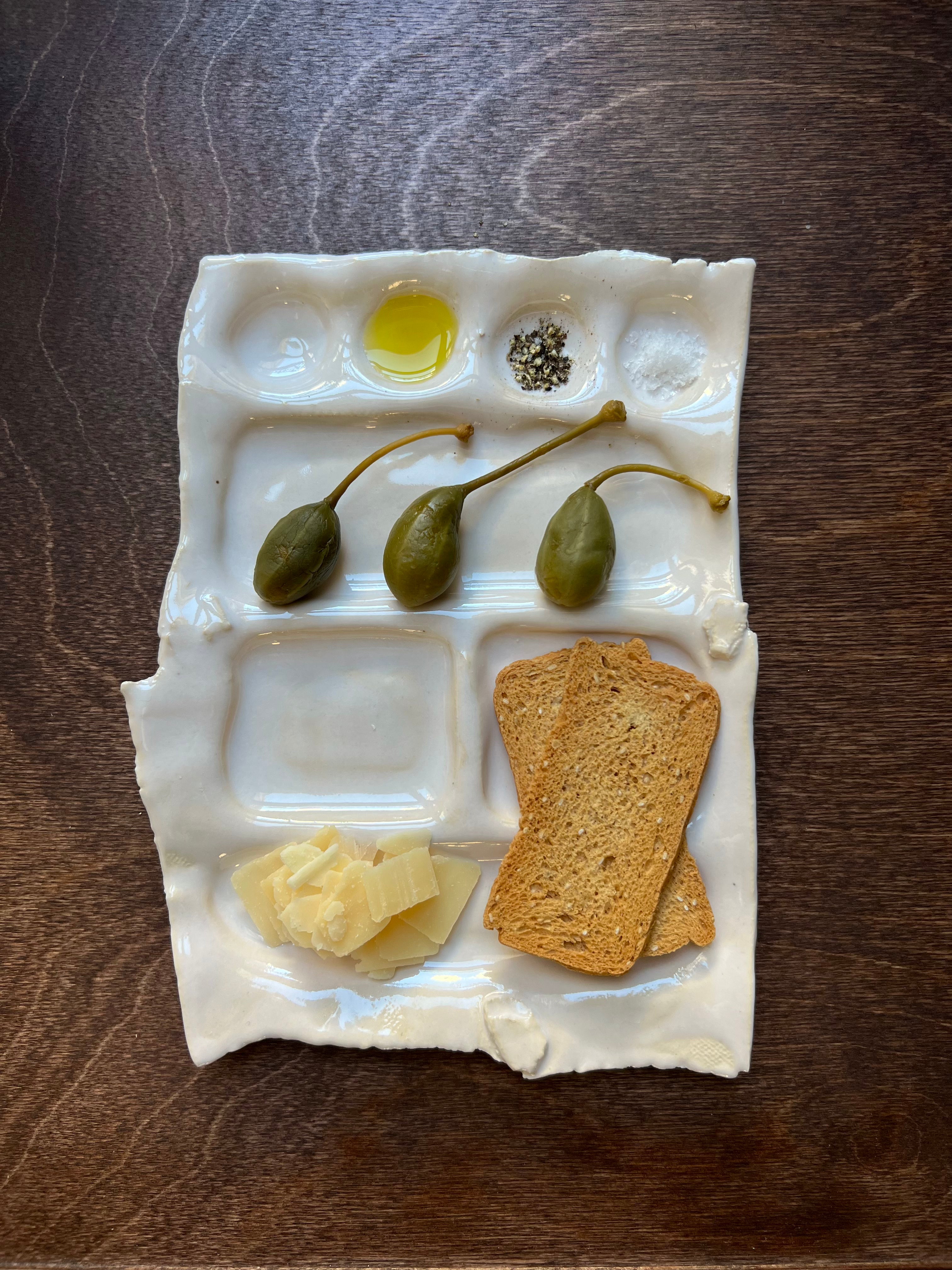 A ceramic Snack Plate from Somthings with compartments containing olive oil, salt and pepper, caperberries, butter slices, and a slice of toasted bread, displayed on a wooden table. The tray features a shiny white.