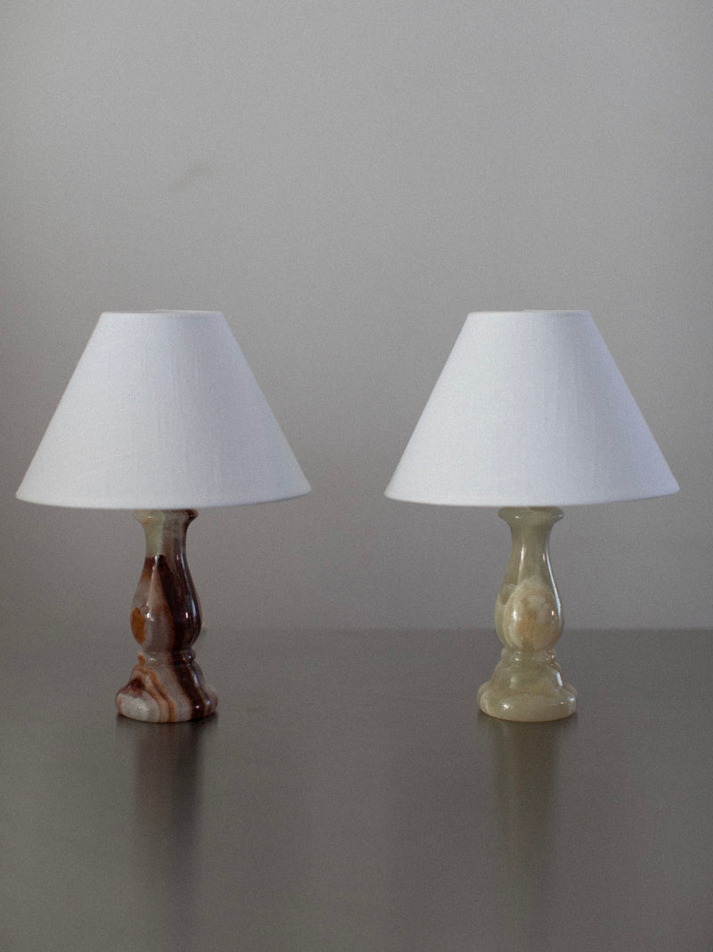 Two small table lamps from the Marble Table Lamps Duo by Out For Lunch are placed side by side on a gray surface. The lamp on the left has an onyx marble base, while the one on the right features a lighter, beige-toned base. Both lamps stand unlit against a plain gray background.