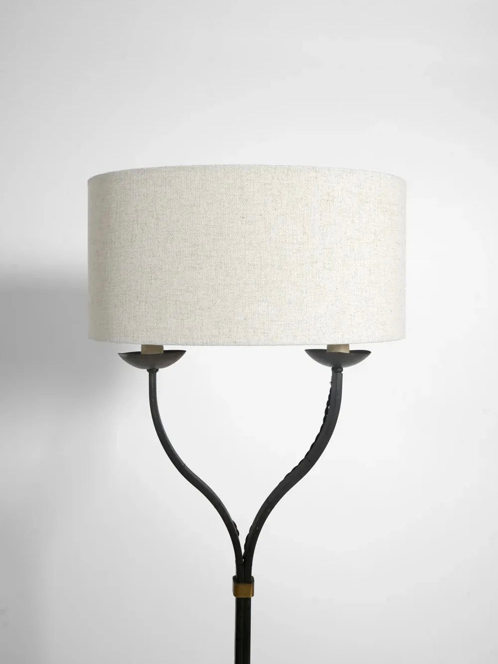 The Iron Floor Lamp from Barracuda Interiors exudes elegance with its white, cylindrical lampshade and black, Y-shaped split stand. The two small cups supporting the shade against a plain white background are reminiscent of 1960s French design.