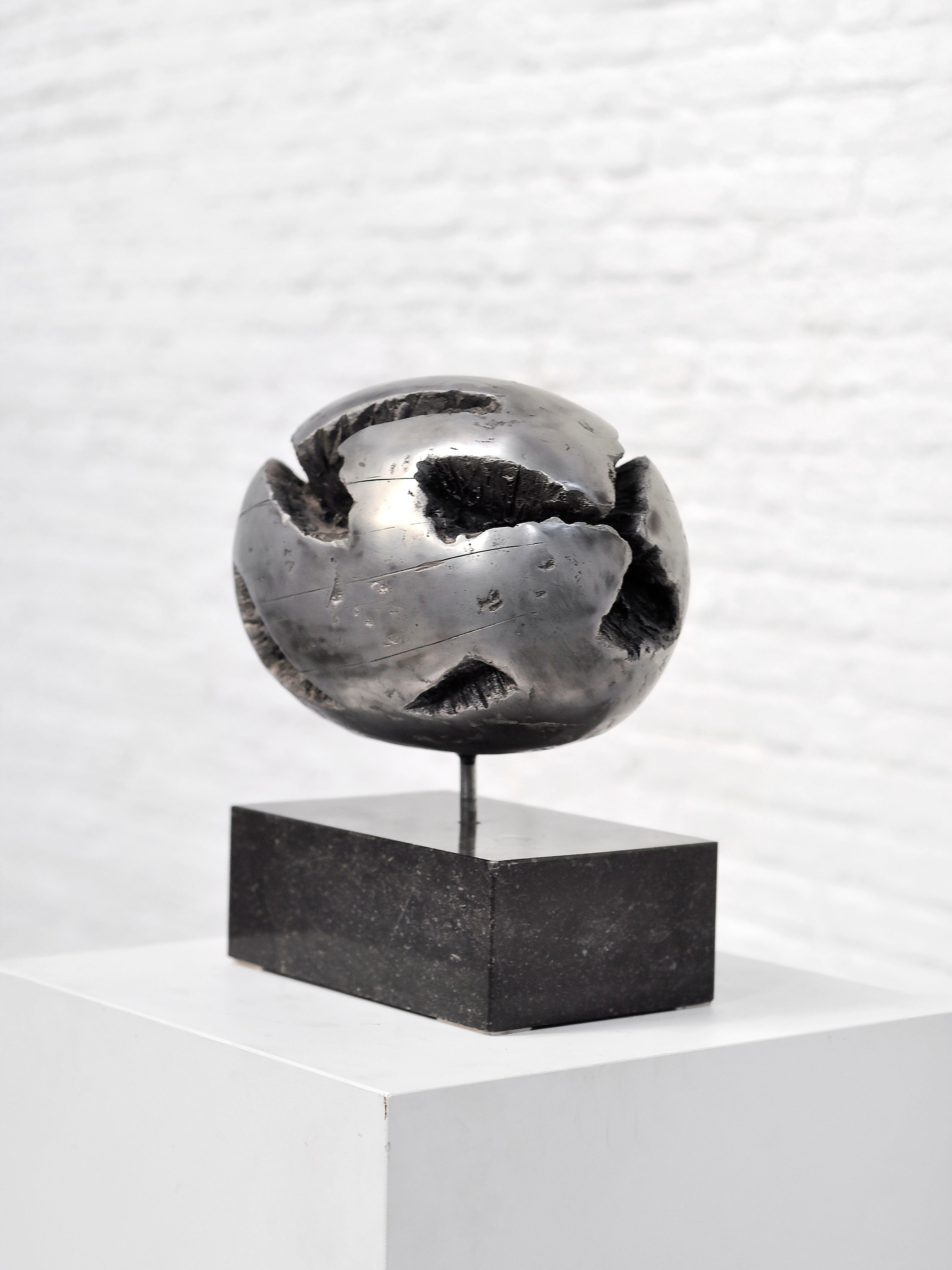 The Introverso 1970's Brutalist Spherical Metal Sculpture, a polished abstract piece with irregular openings on a black rectangular base, is showcased on a white pedestal against a light brick wall, capturing the raw materials and aesthetics of the era.