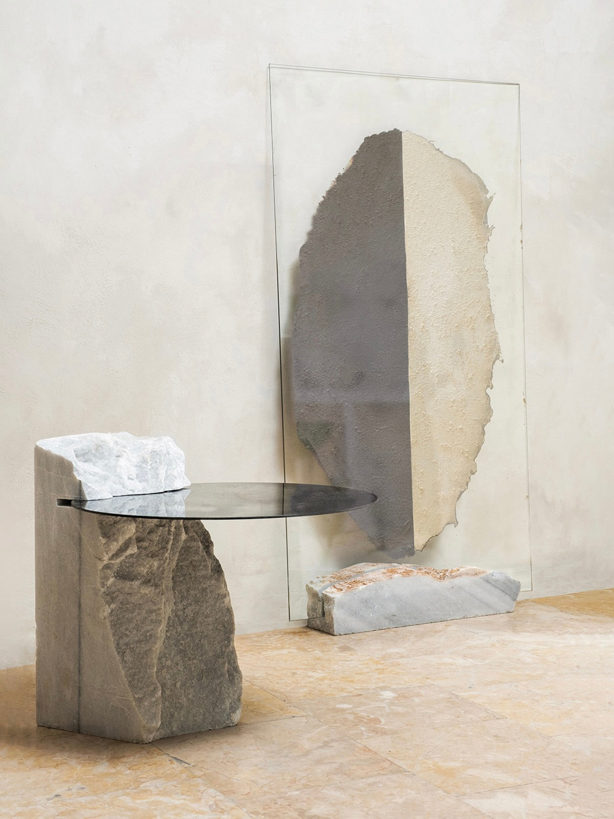 The Cloud Mirror Side Table by HOMA showcases a glass top on irregular stone slabs. Background elements include oxidized silver mirrors with textured, earth-toned stones against a light-colored wall.