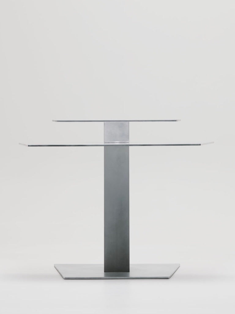 The Beam Desk by Marquel Williams is a sleek, modern piece featuring a transparent glass top and an I-beam-inspired metal pedestal base. Its handcrafted design highlights clean lines and simplicity, all set against a plain white background.