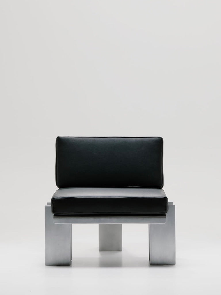 The Beam Lounge Chair, crafted by Marquel Williams, showcases a minimalist design with a sleek black cushion and sturdy silver metal base. It features luxurious full-grain leather against a light backdrop for a modern aesthetic.