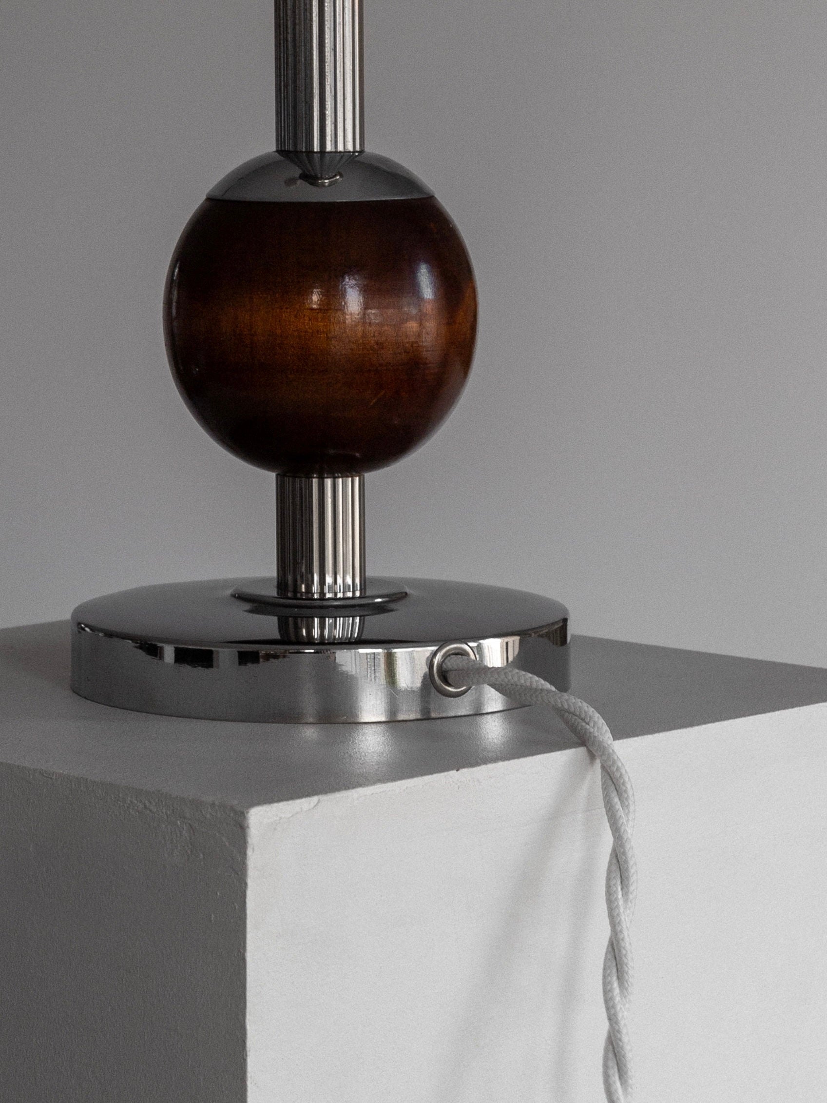 Displayed on a white pedestal, the Wood & Chrome Table Lamp by Spigel exudes 1930s Swedish Art Deco charm. It showcases a gleaming chrome and wood base, adorned with a polished wooden spherical accent and a twisted white cord extending from it. The setup is set against a simple gray wall backdrop.