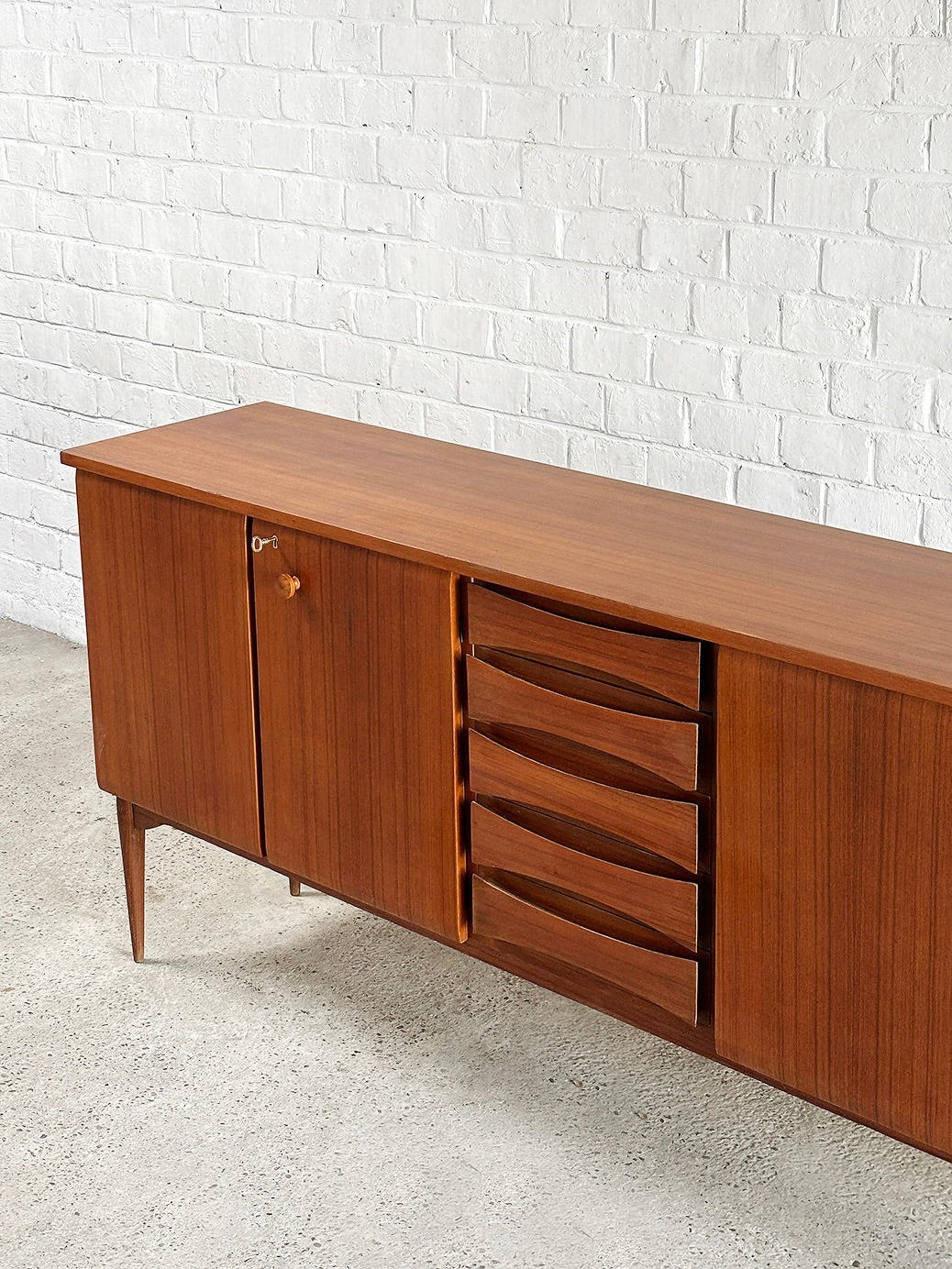 An Introverso Italian Modernist Teak Sideboard by Vittorio Dassi from the 1960s enhances a white brick wall. With sleek, minimalist lines, it features a cabinet door on the left and five woodgrain-finished pull-out drawers on the right, embodying mid-century modern style.